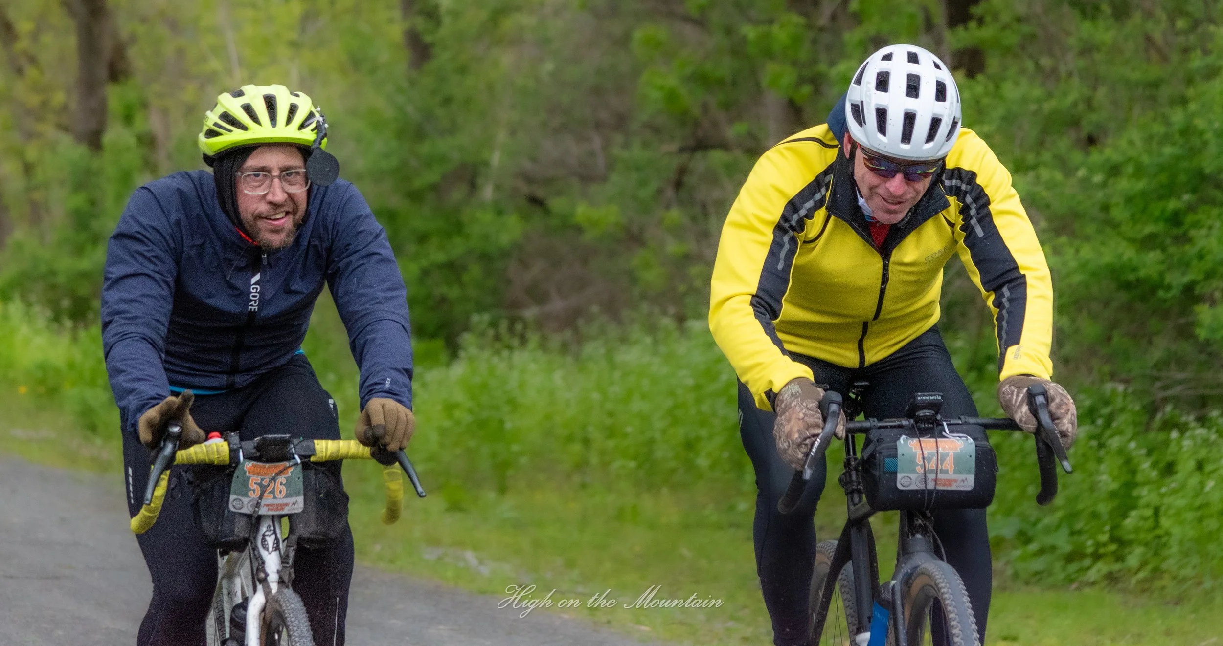 Two men riding mountain bikes on a trail through a green forest, wearing helmets and outdoor gear.