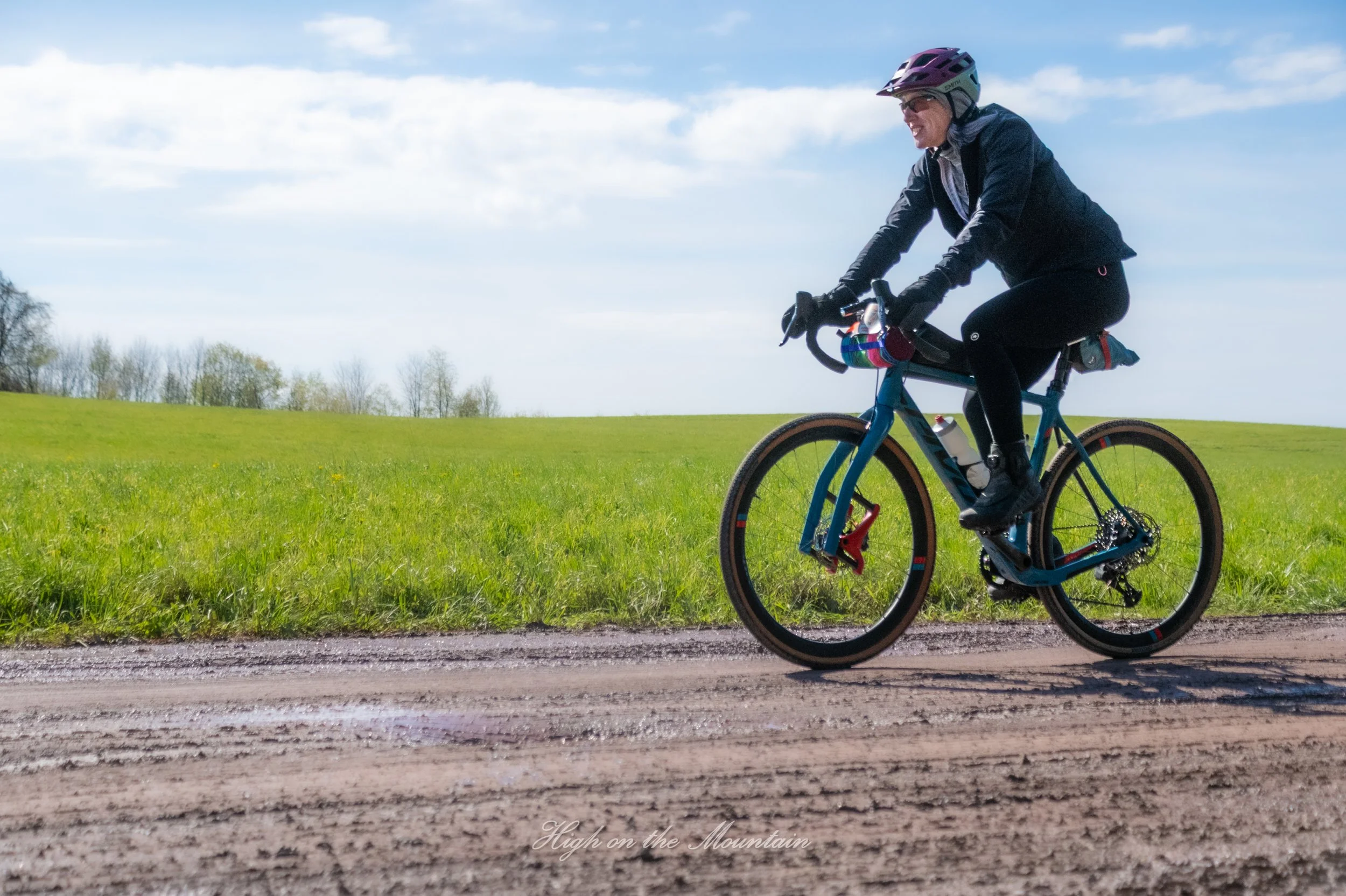 Woman riding a blue mountain bike on a dirt trail in a green field with trees and cloudy sky in the background.