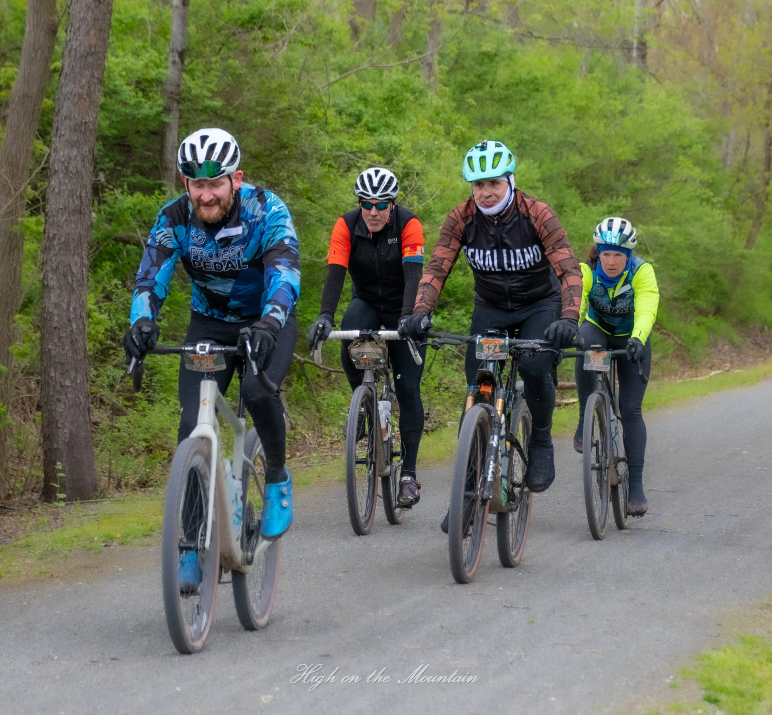 Four cyclists riding mountain bikes on a trail surrounded by green trees. They are wearing helmets and sports gear.