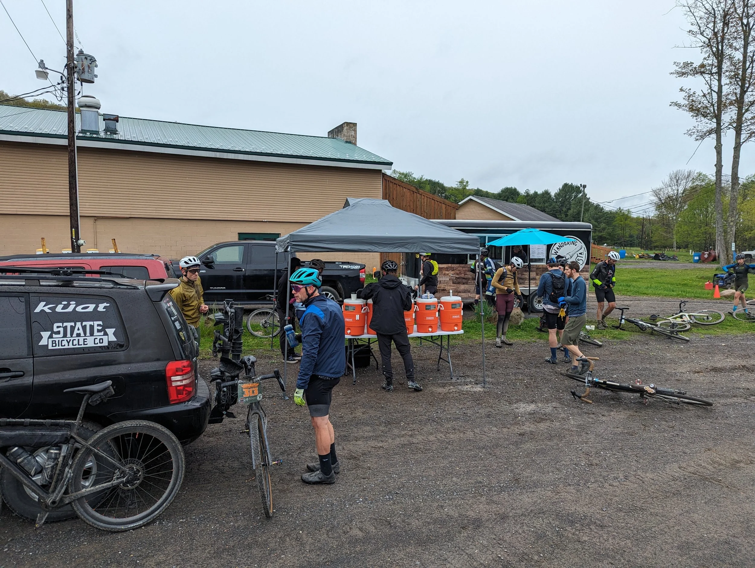 People gathering for a mountain biking event at a registration table outside a building on a cloudy day. Bicycles are on the ground, and some individuals are wearing helmets and cycling gear.
