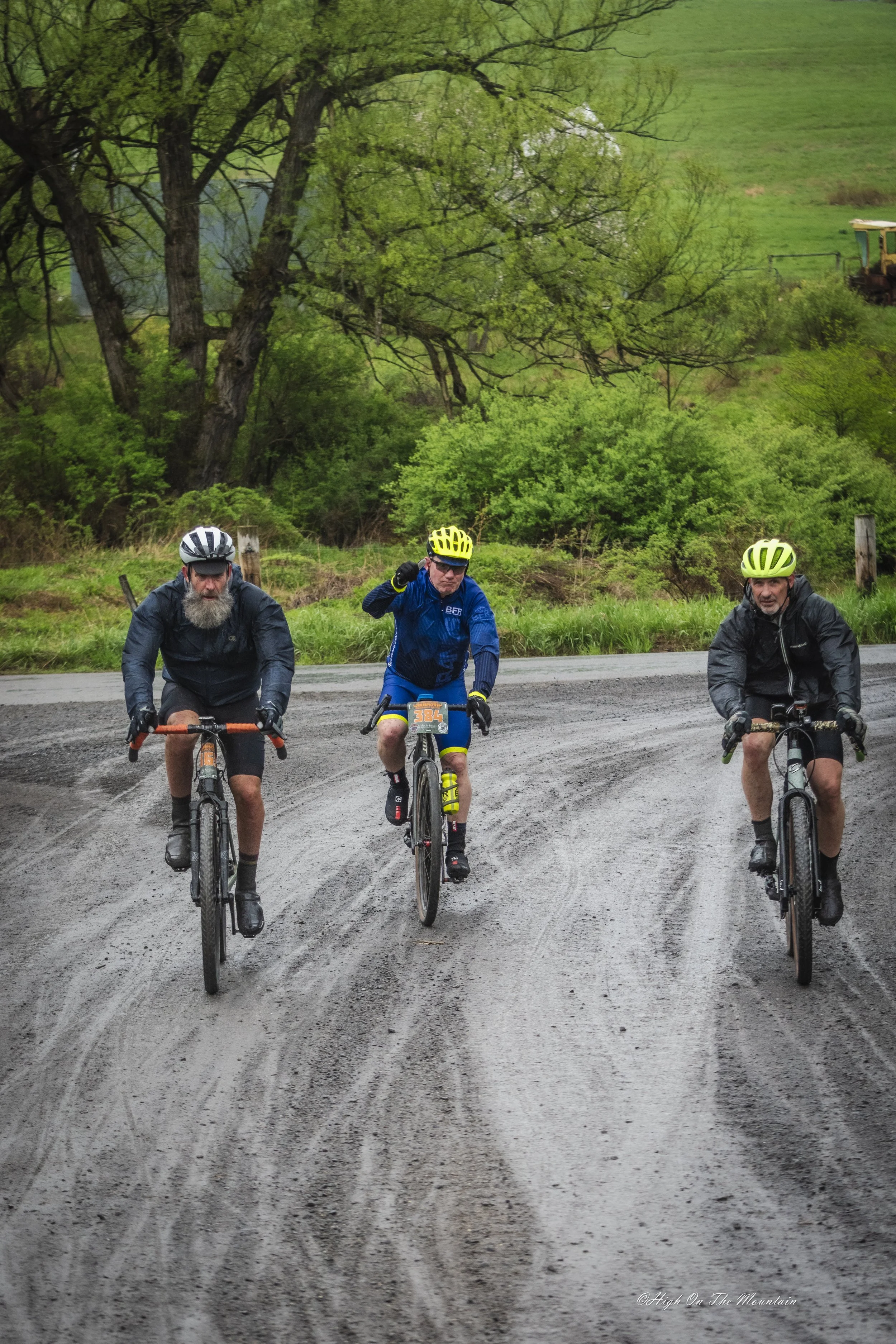 Three men riding mountain bikes on a dirt trail surrounded by green trees and grass, with a hill and some farm equipment in the background.