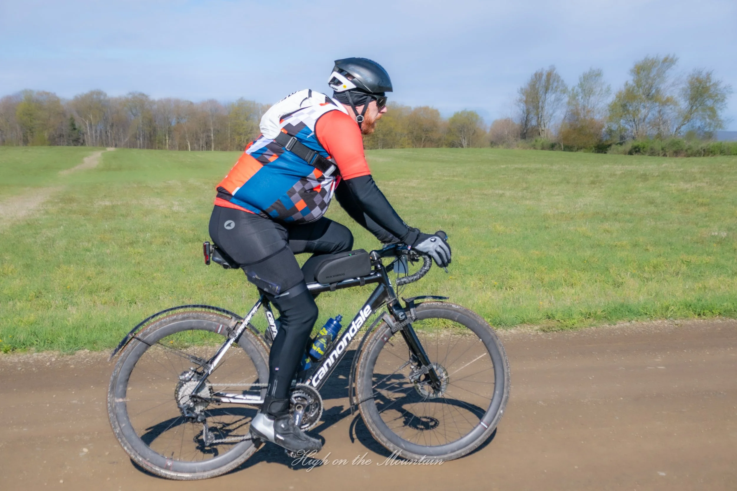 A man riding a mountain bike on a dirt trail in a grassy open field with trees in the background, wearing a helmet, sunglasses, a red and black cycling outfit, and black gloves.