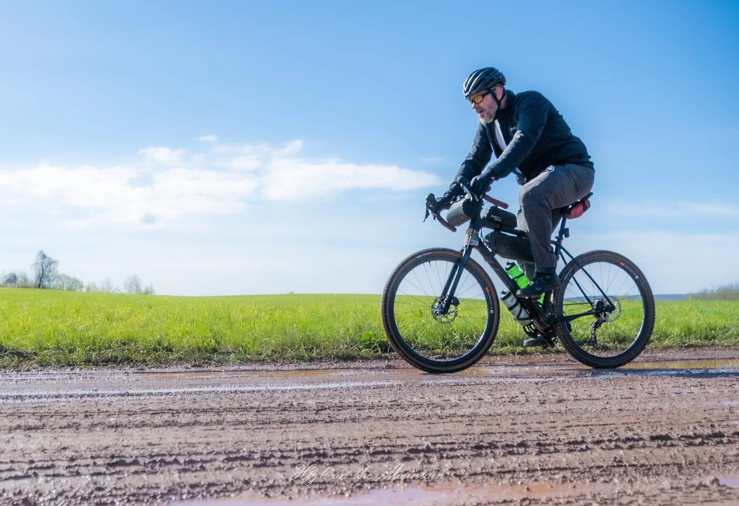 A man wearing a black helmet, sunglasses, and dark athletic clothing riding a mountain bike on a dirt trail in a grassy field with trees in the distance under a blue sky with some clouds.