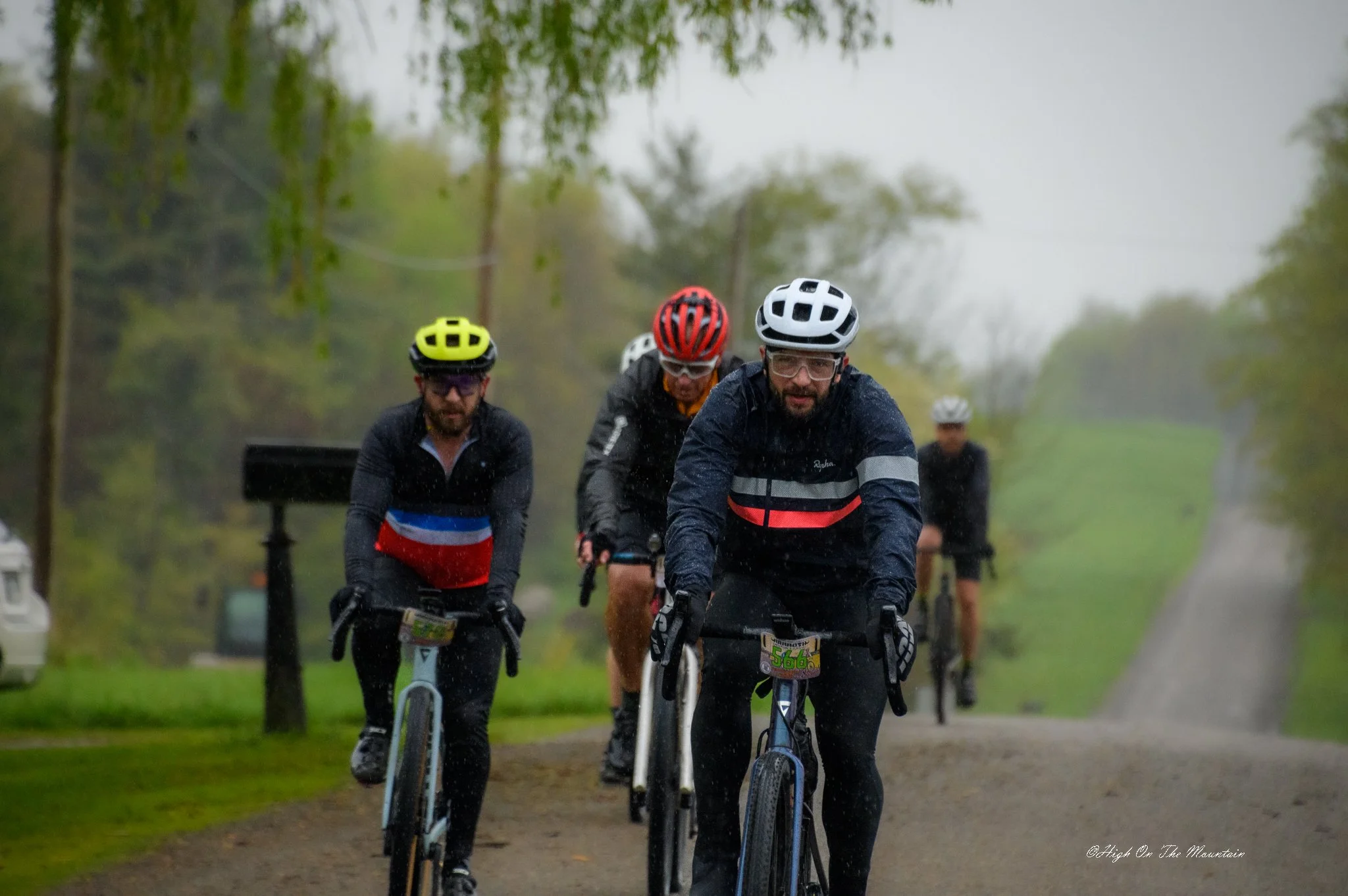 Four cyclists riding on a wet, muddy trail in rainy weather, wearing helmets and rain gear, with trees and a cloudy sky in the background.