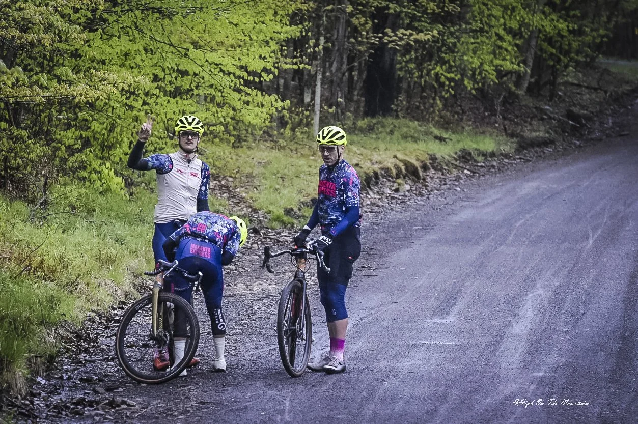 Three mountain bikers wearing colorful cycling gear and helmets, standing on a gravel trail surrounded by green trees, with one waving at the camera.