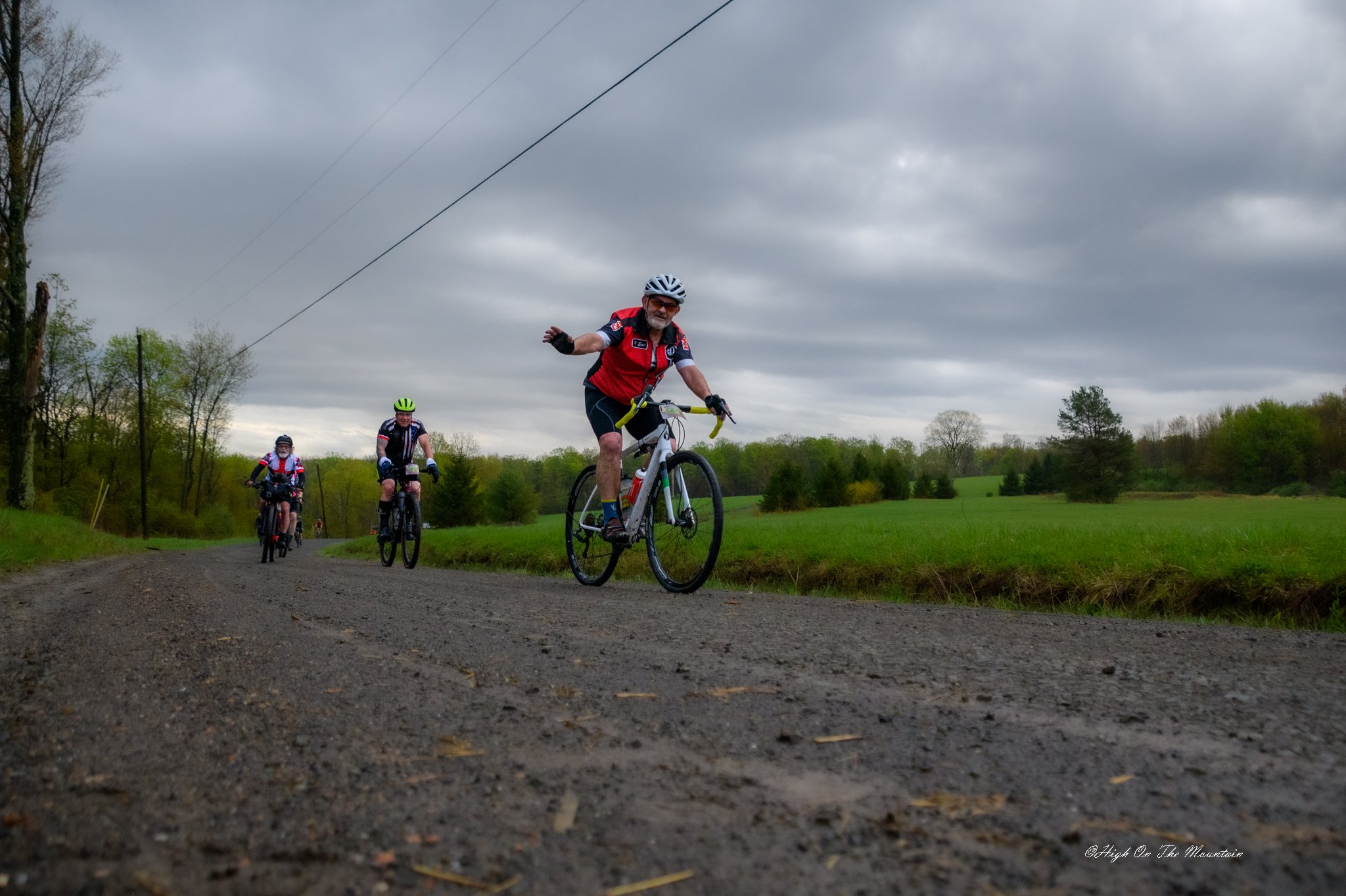 Three cyclists riding on a dirt road in a rural area under a cloudy sky, surrounded by green fields and trees.