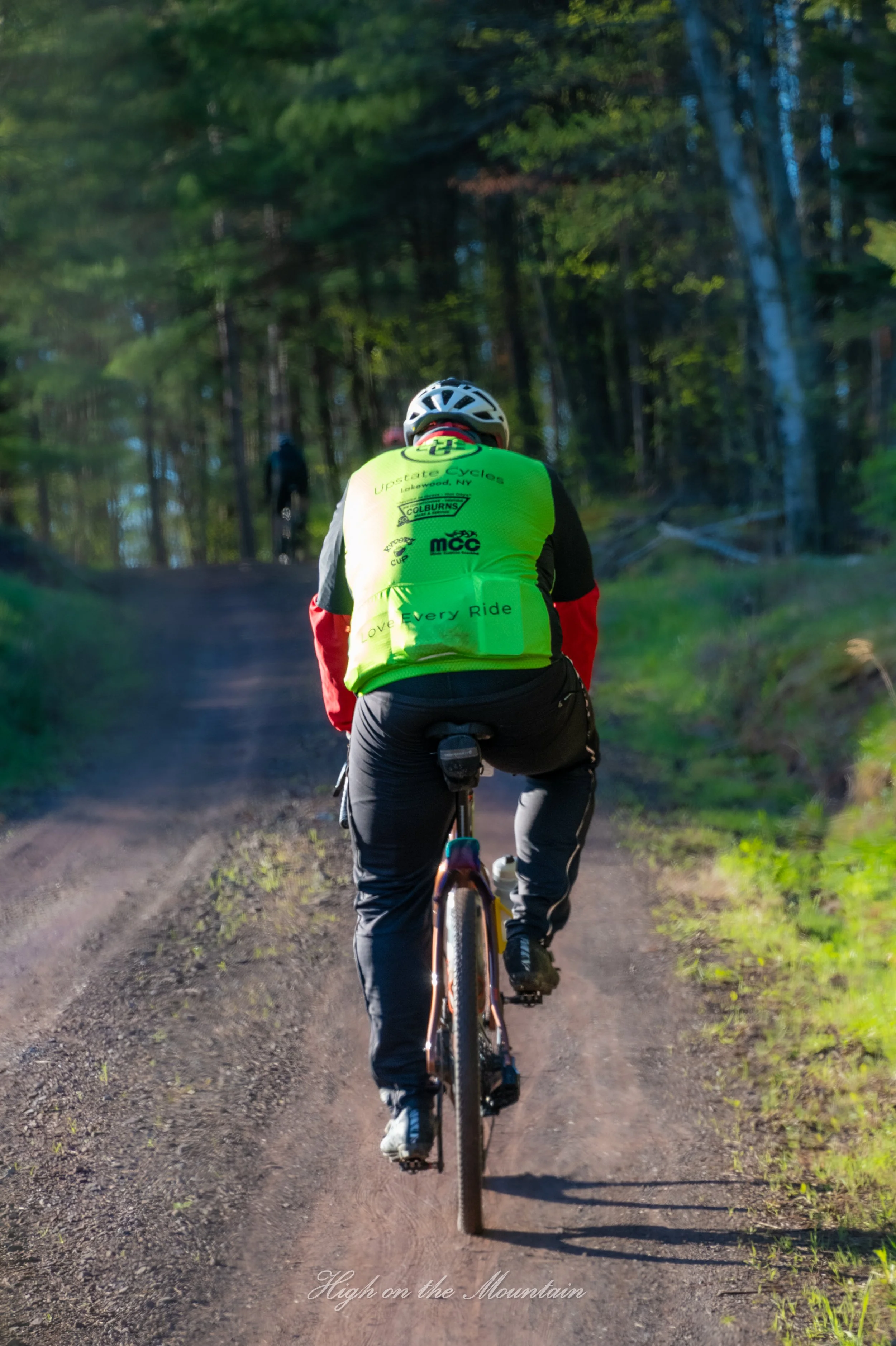 A person riding a mountain bike on a dirt trail in the woods, wearing a bright green vest with various logos and the words "Love Every Ride," with trees and sunlight in the background.