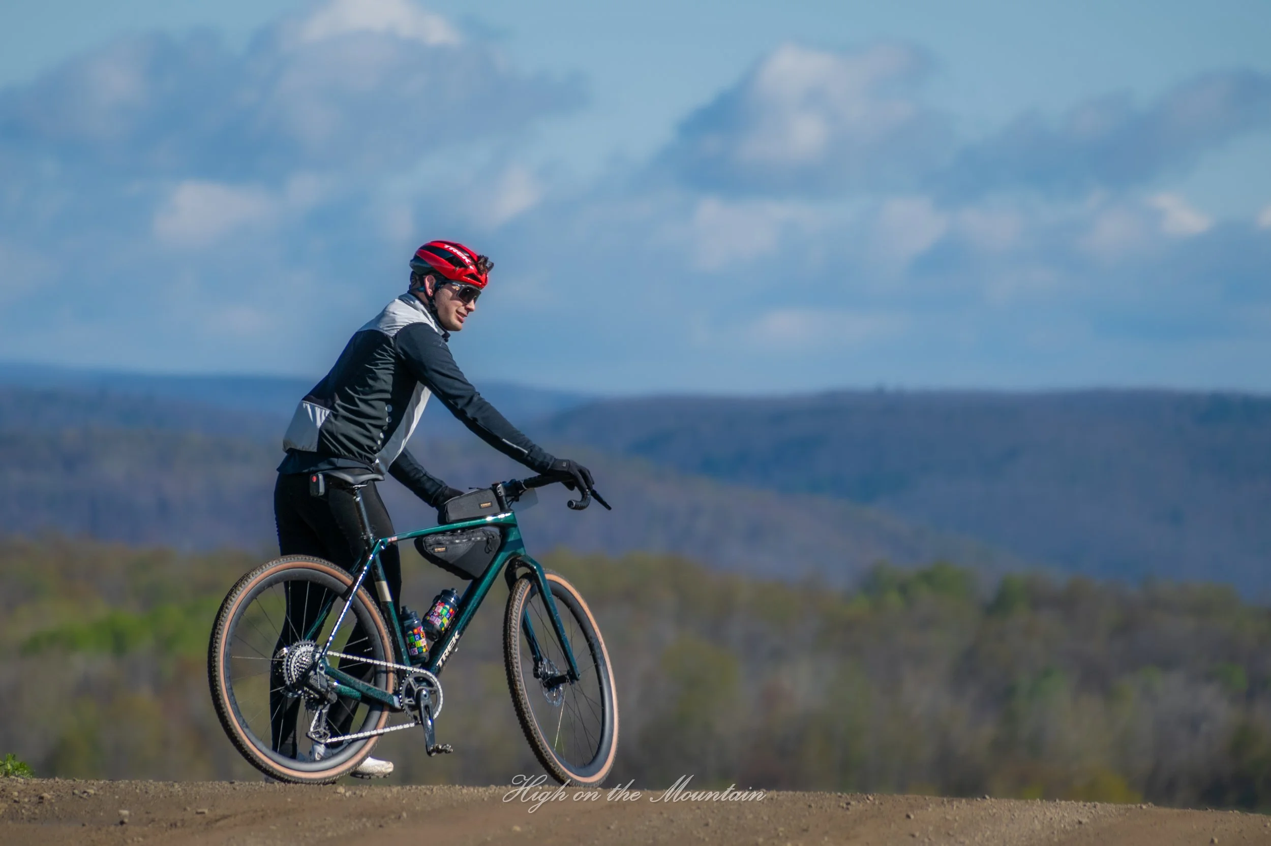 A person wearing a red helmet, sunglasses, and black and gray athletic clothing riding a mountain bike on a trail with a blurred background of mountains and sky.