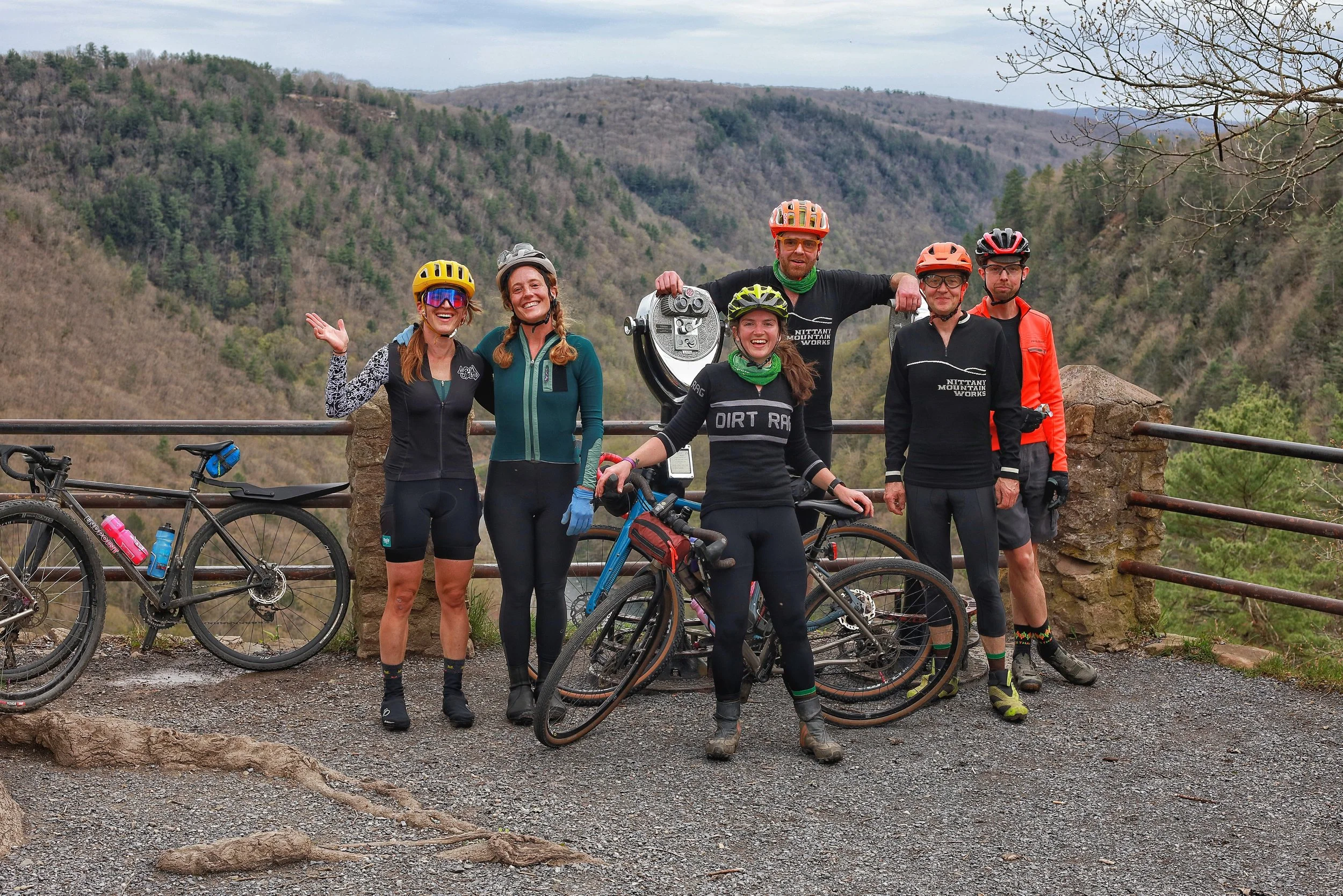 Group of six cyclists in mountain biking gear posing with their bikes on a scenic overlook with mountainous terrain in the background.