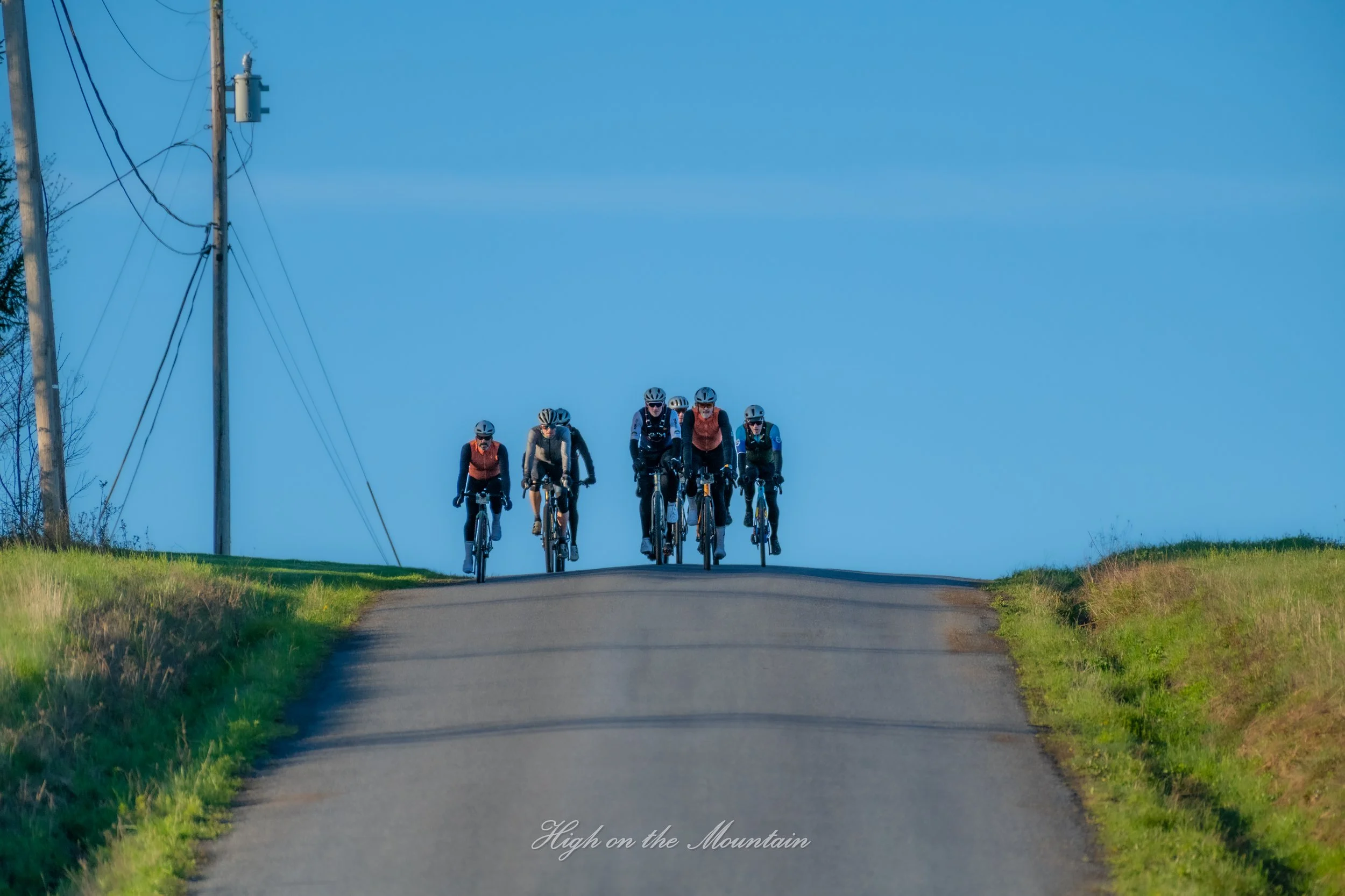 Cyclists riding on a mountain trail with a clear blue sky and power lines overhead.