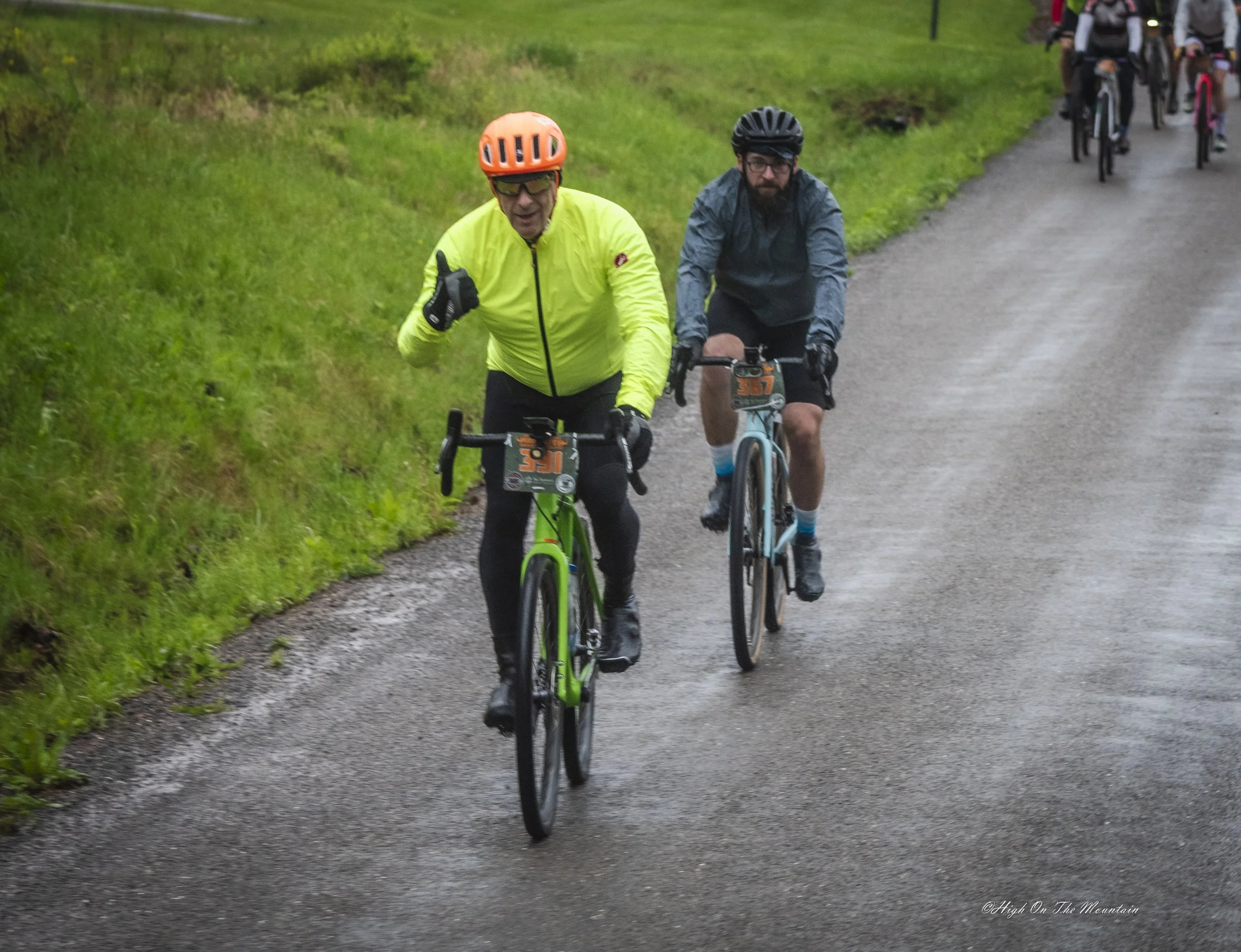 Two men riding bikes on a gravel path surrounded by green grass and foliage, with a group of cyclists in the background.