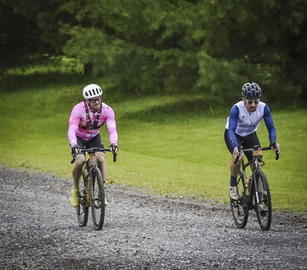 Two male cyclists riding on a gravel trail through a green park with trees in the background. One is wearing a pink jacket and white helmet, the other is in a blue and white jersey with a black helmet.
