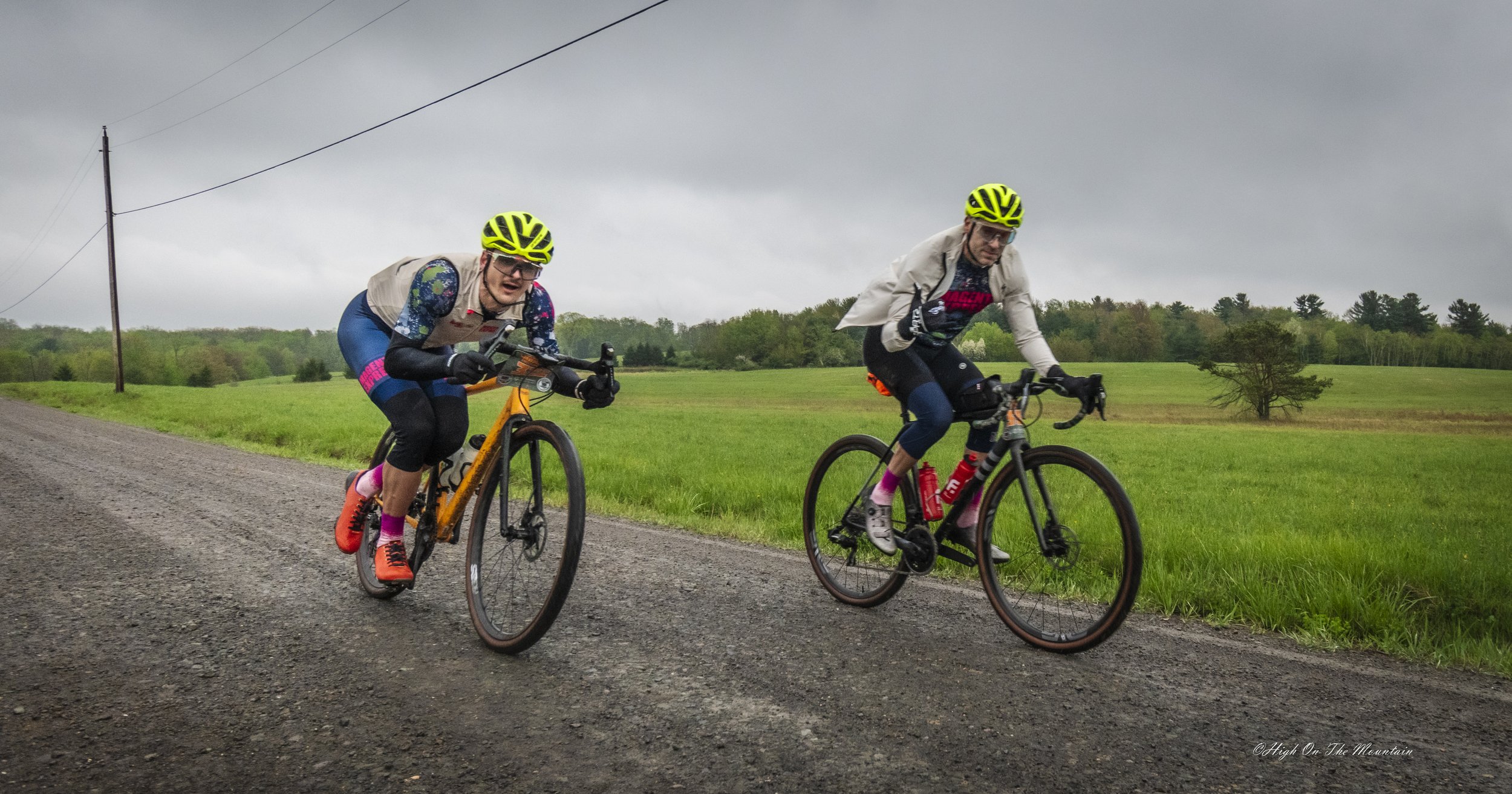 Two cyclists riding on a gravel road in a rural area with green fields and trees under a cloudy sky.