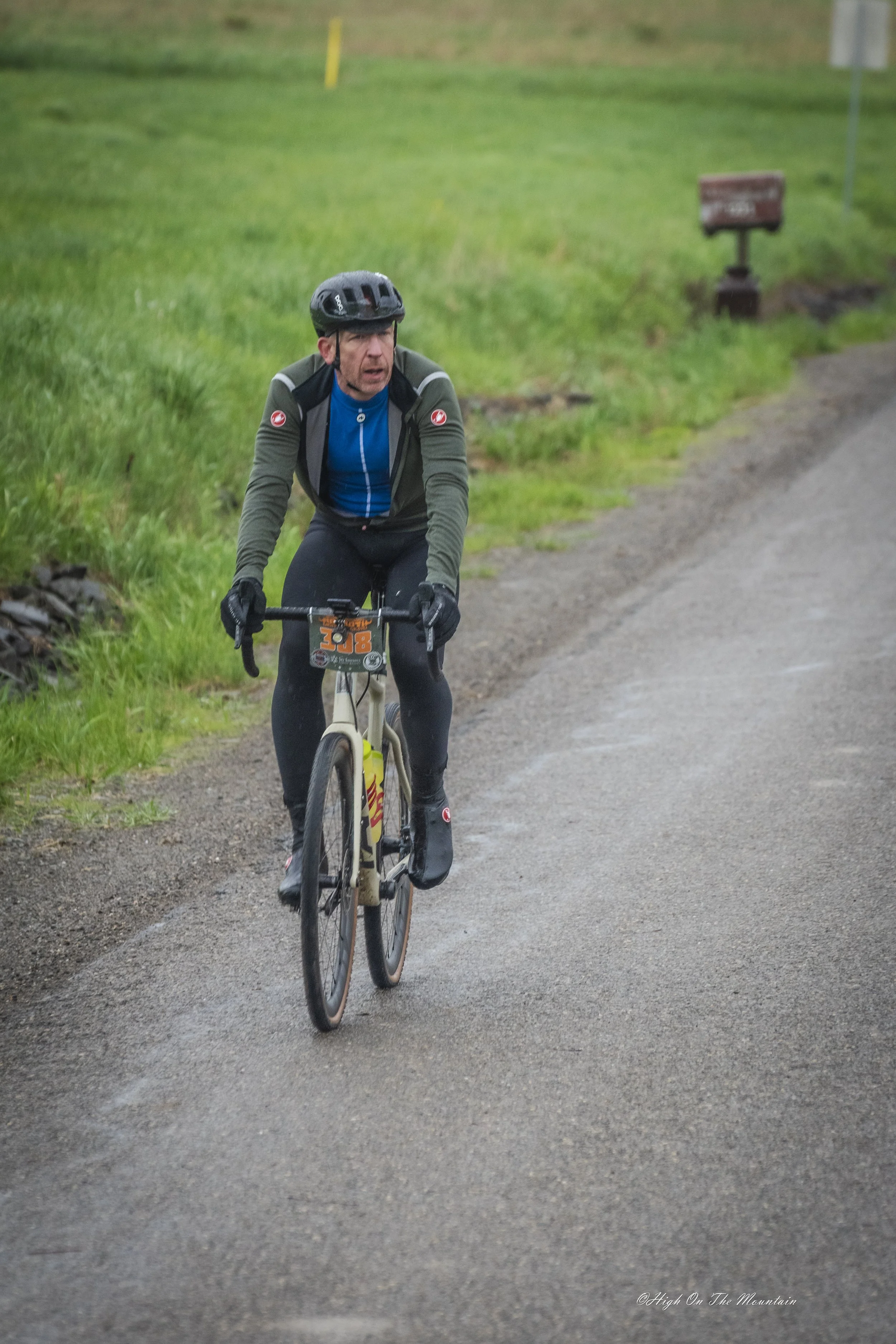 A man wearing a black helmet and green outdoor jacket rides a mountain bike on a dirt path. The background shows green grass and a few yellow poles. He appears focused and is riding on a trail through a natural area.