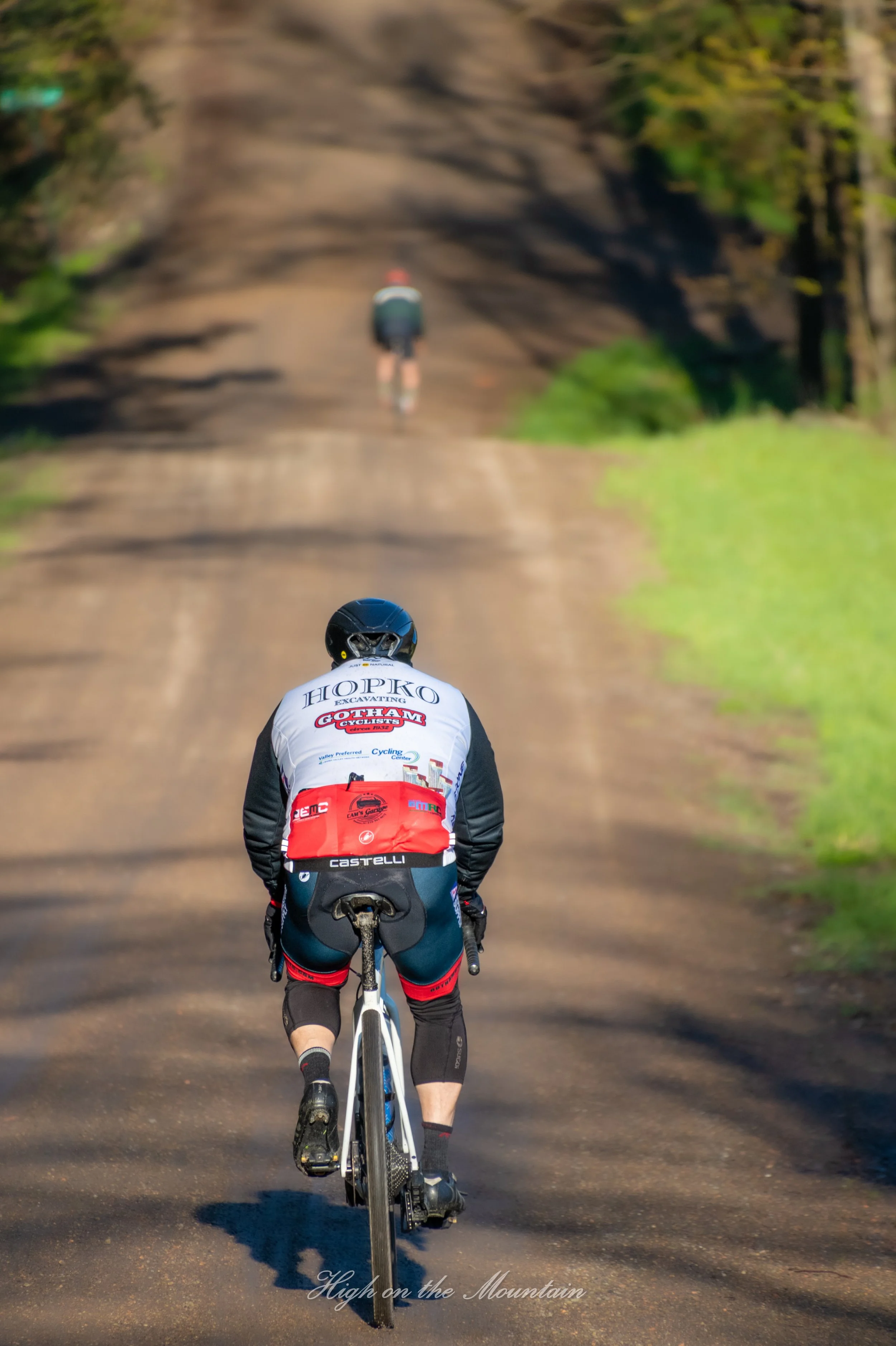 A cyclist riding on a dirt trail in a wooded area with trees on both sides, seen from behind, wearing a cycling helmet and a jersey with sponsored logos. The trail extends into the distance with a gentle incline. Text at the bottom reads "High on the