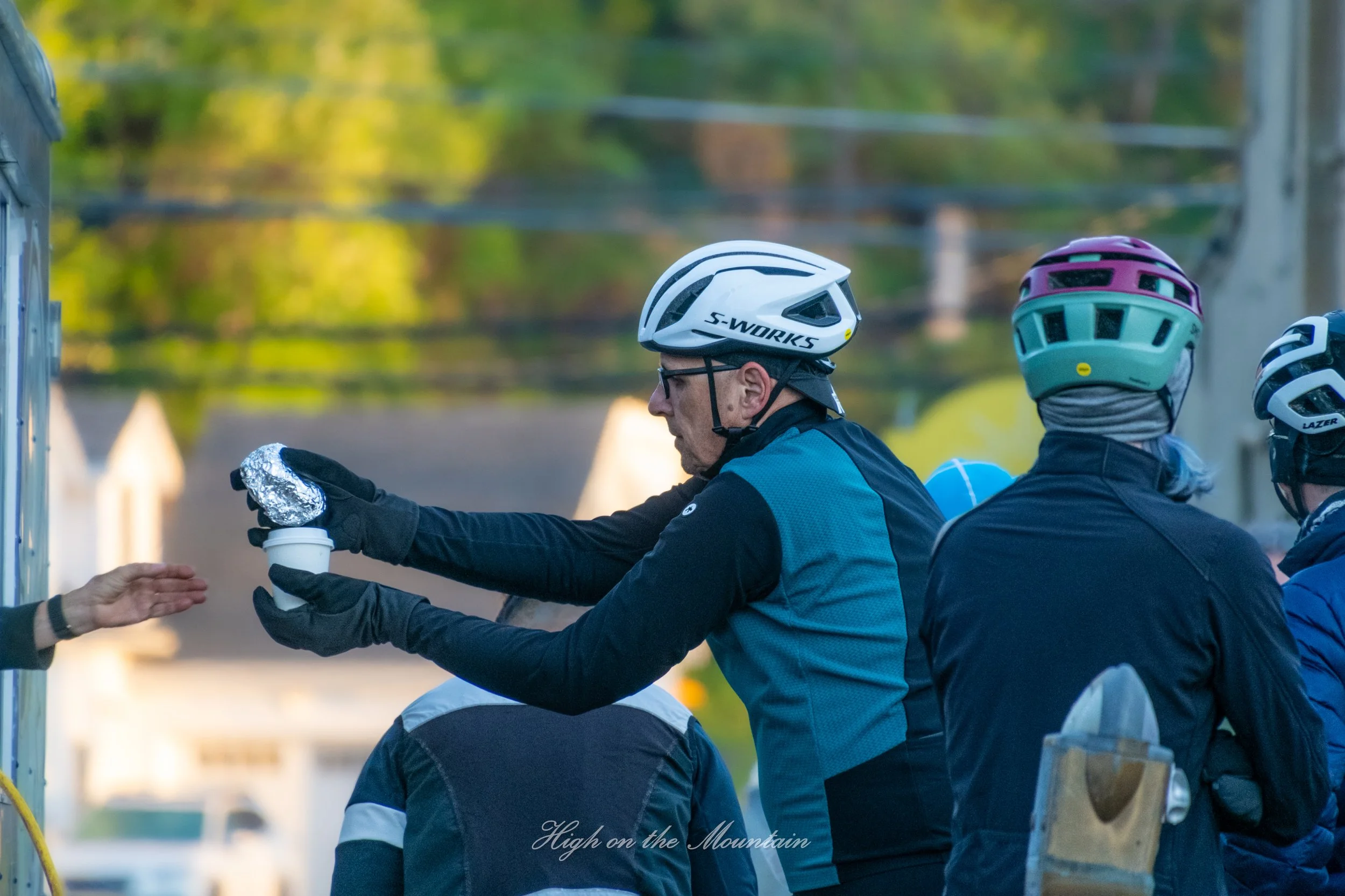A man in a helmet and outdoor gear is handing a paper cup to another person outdoors during daytime. There are other people wearing helmets nearby. The background features trees and blurred houses, indicating a neighborhood setting.