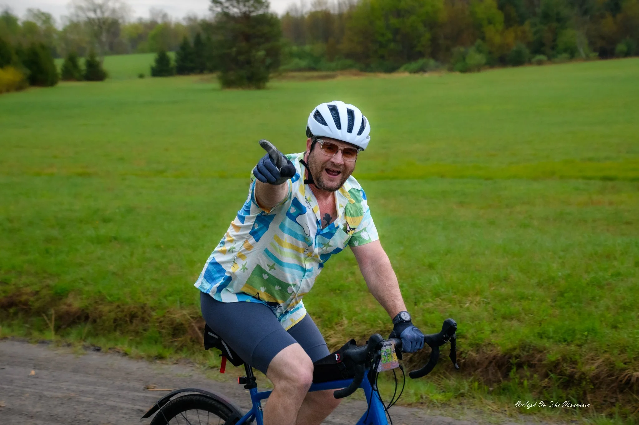 A man wearing a white bike helmet and glasses is riding a blue bicycle on a trail through a grassy area with trees in the background. He is smiling and pointing with his right hand.