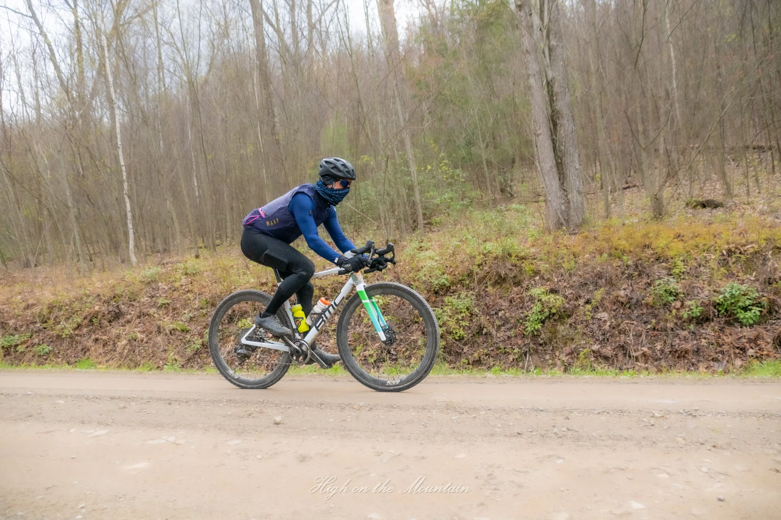 Person wearing a black helmet and face covering riding a mountain bike on a dirt path through a forest with leafless trees.