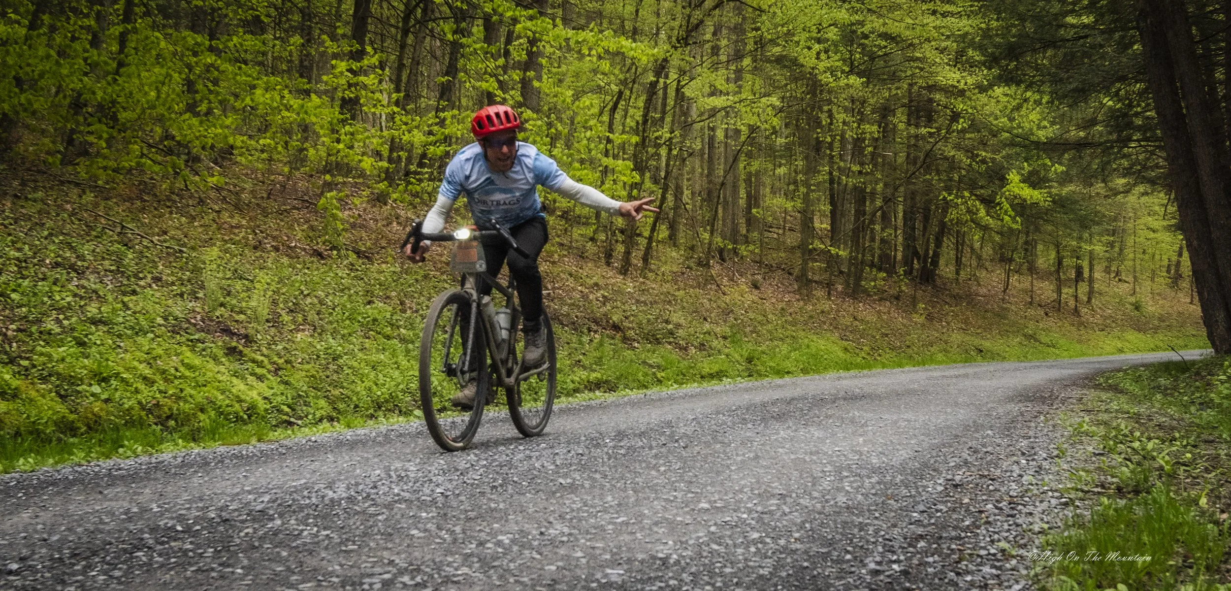 A person riding a mountain bike on a gravel trail through a forest with green foliage, wearing a red helmet and a light blue cycling jersey, making a peace sign with their right hand.