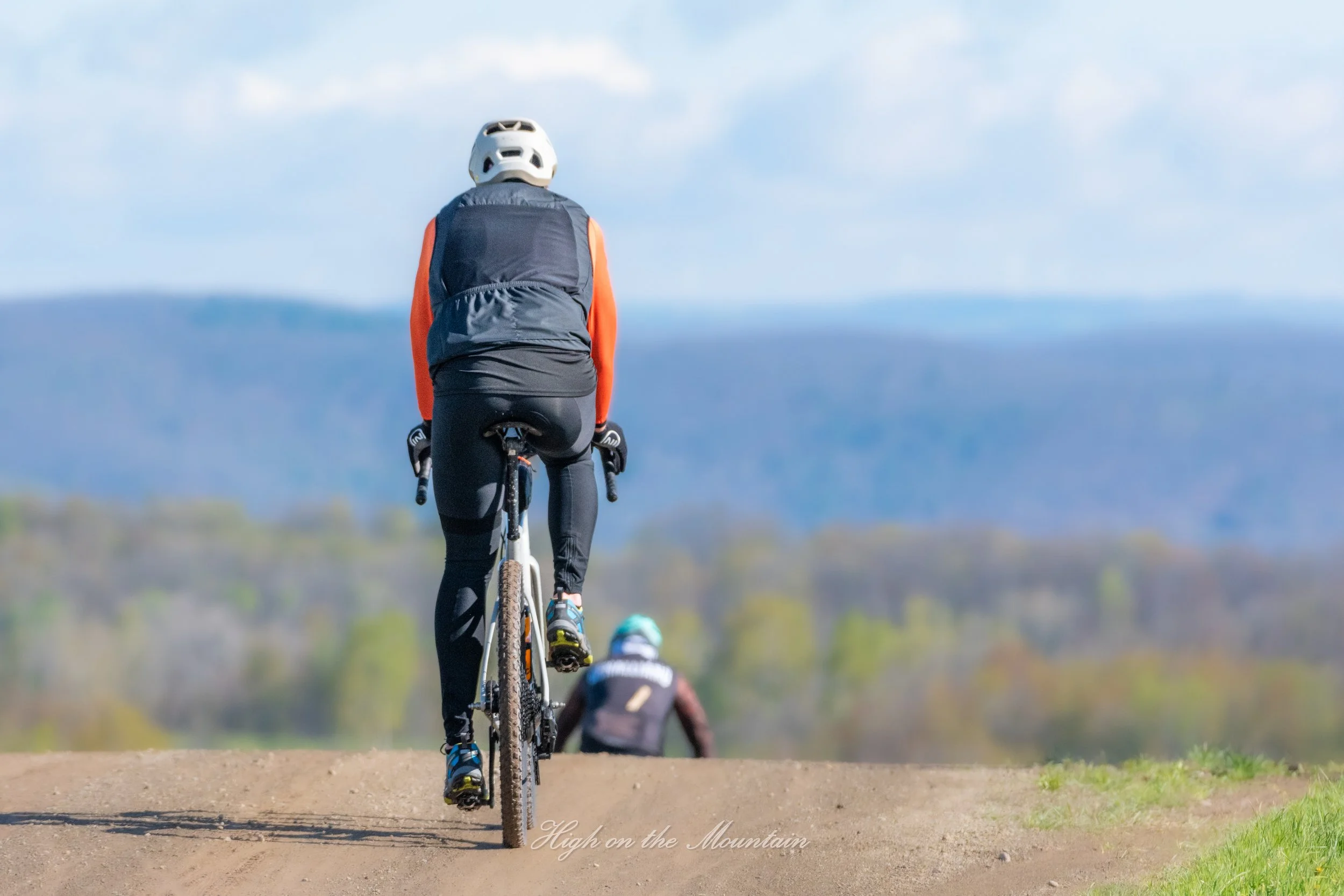 A person wearing a white helmet and a dark vest riding a mountain bike on a dirt trail with mountains and blue sky in the background.