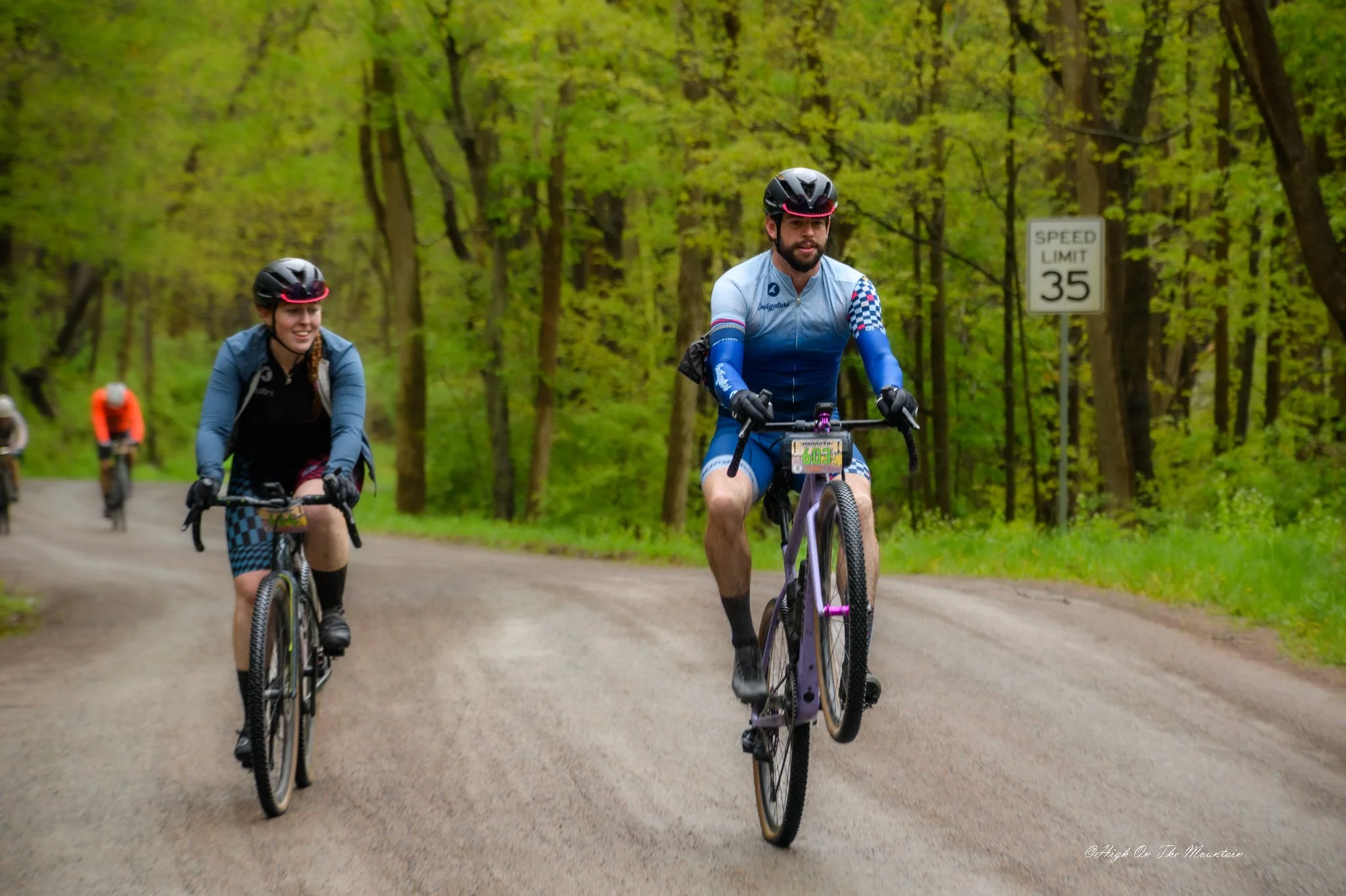 Three cyclists riding on a dirt trail through a green forest, with speed limit sign in background.