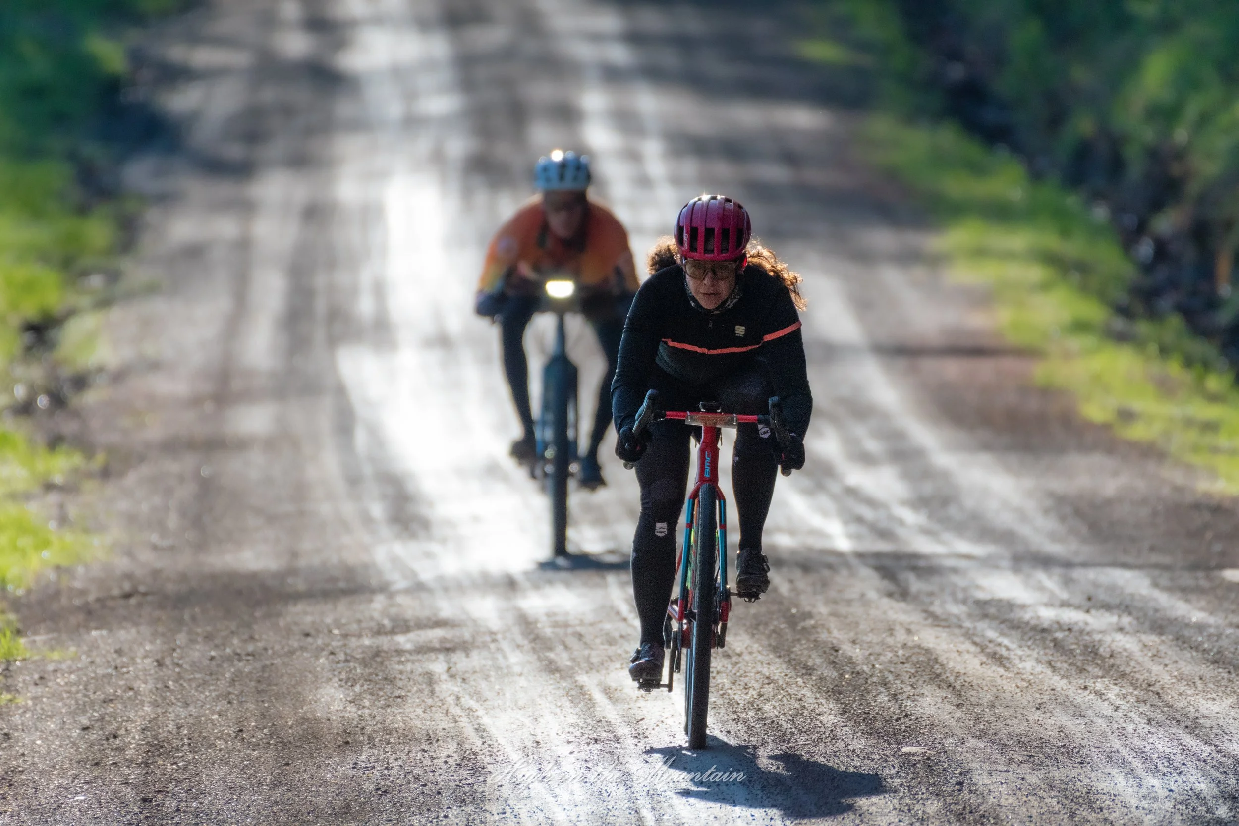 Two women riding bicycles on a dirt forest trail, wearing helmets and athletic clothing, with green trees on both sides of the path.