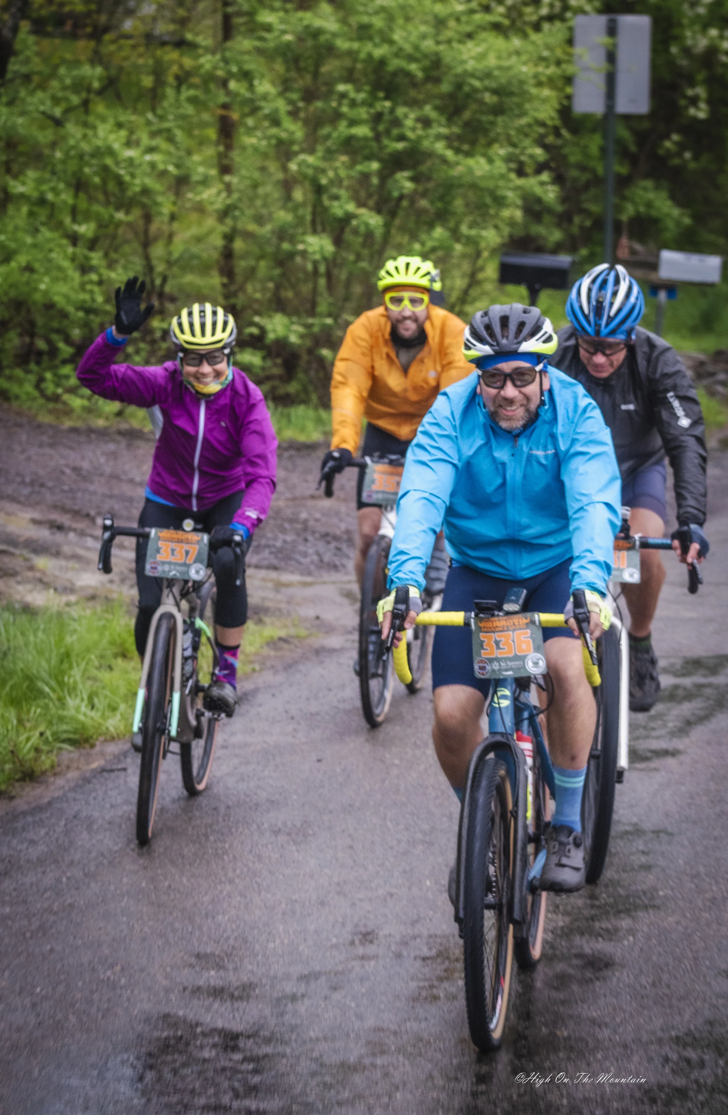 Four cyclists riding on a forest trail, wearing helmets and colorful jackets, some smiling and waving, surrounded by green trees.