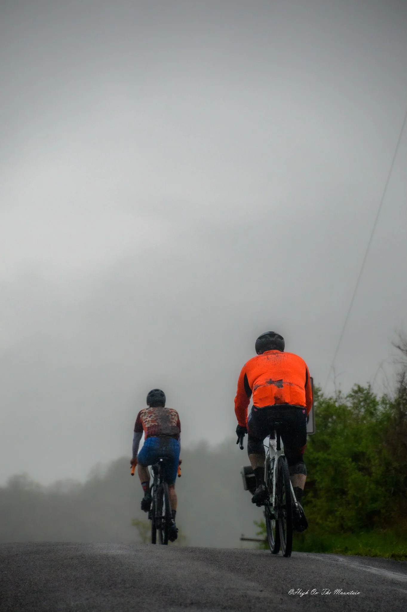 Three cyclists riding on a road with a misty or foggy background, wearing helmets and cycling gear, with trees visible to the right.