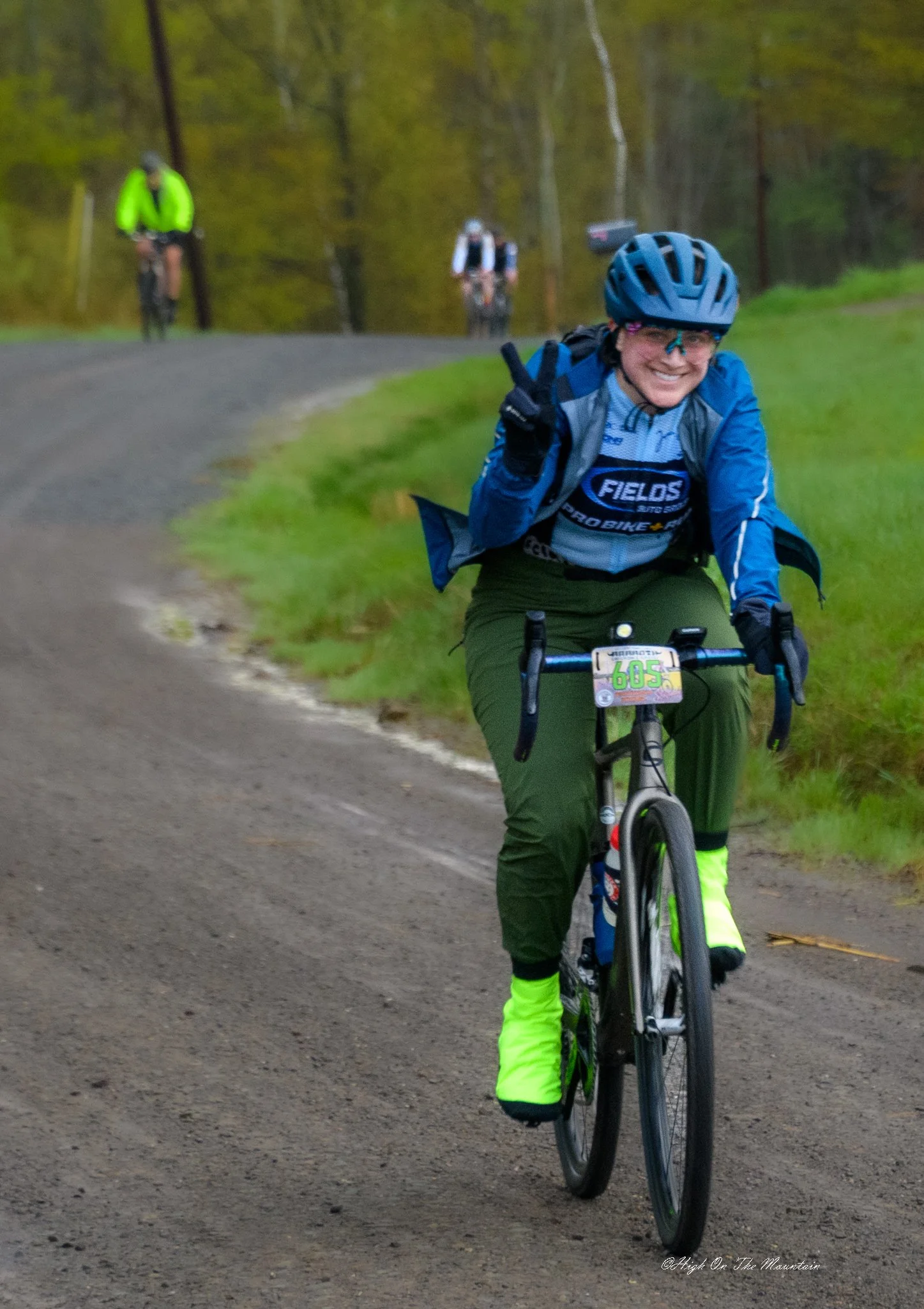 A woman riding a bicycle on a trail in the woods, smiling and wearing a blue helmet, glasses, a blue jacket, and high-visibility pants. There are three other cyclists in the background on the trail, surrounded by trees with green foliage.