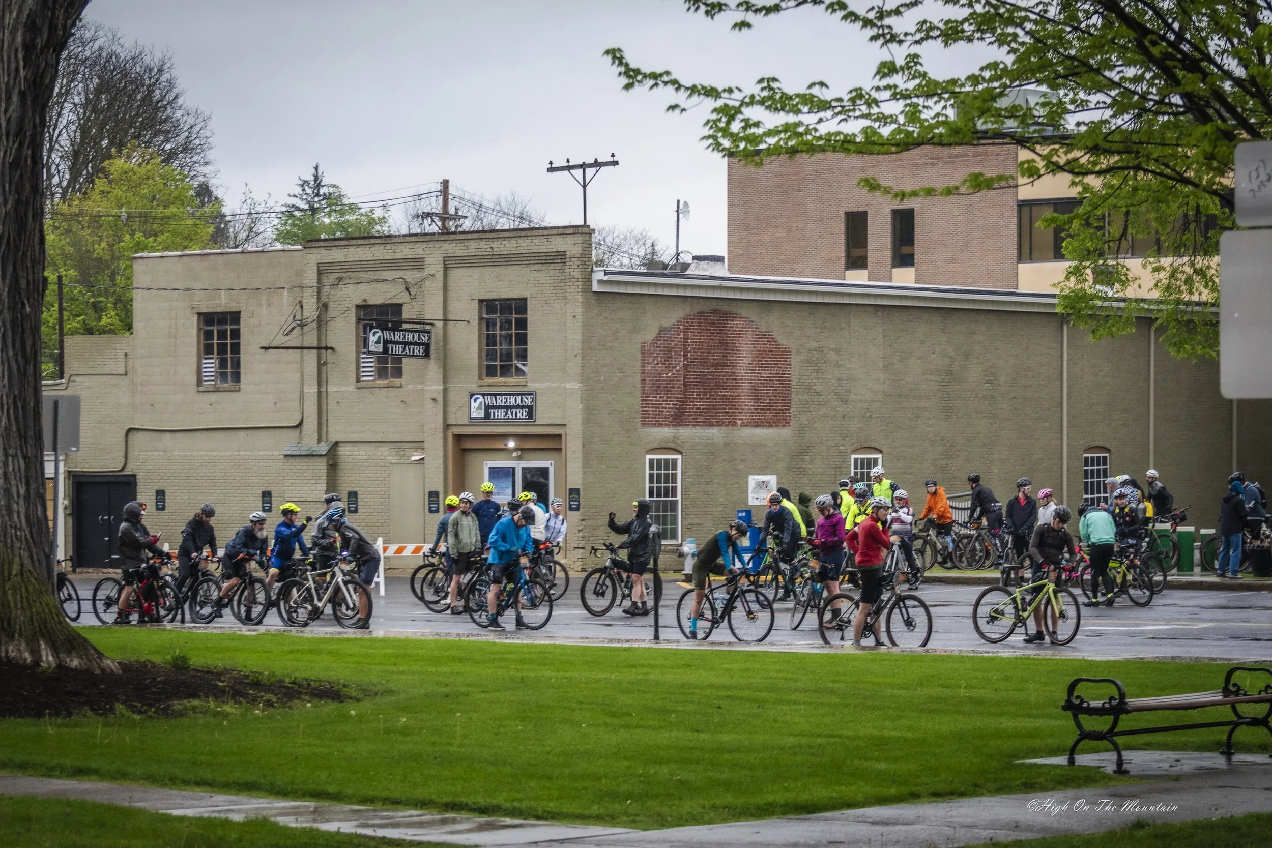 A group of cyclists gathered together outside a building on a cloudy day, surrounded by trees and greenery.
