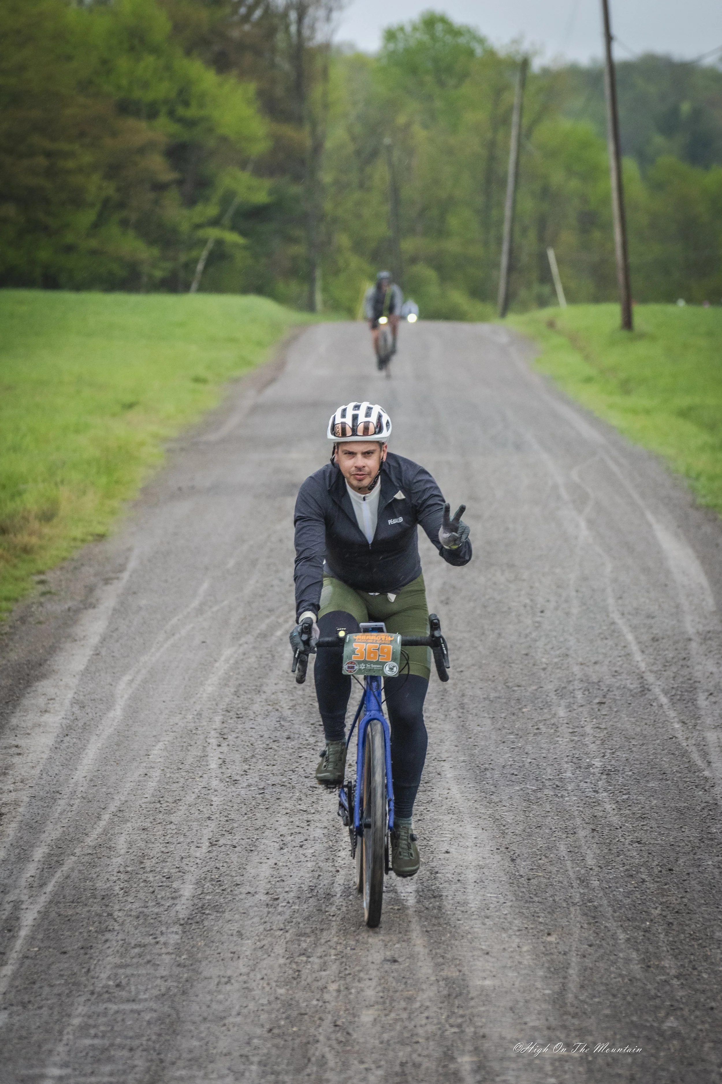 A man cycling on a dirt road with a peace sign, wearing a helmet and cycling gear, with two more cyclists in the background. The scene is outdoors with green grass and trees on either side of the road.