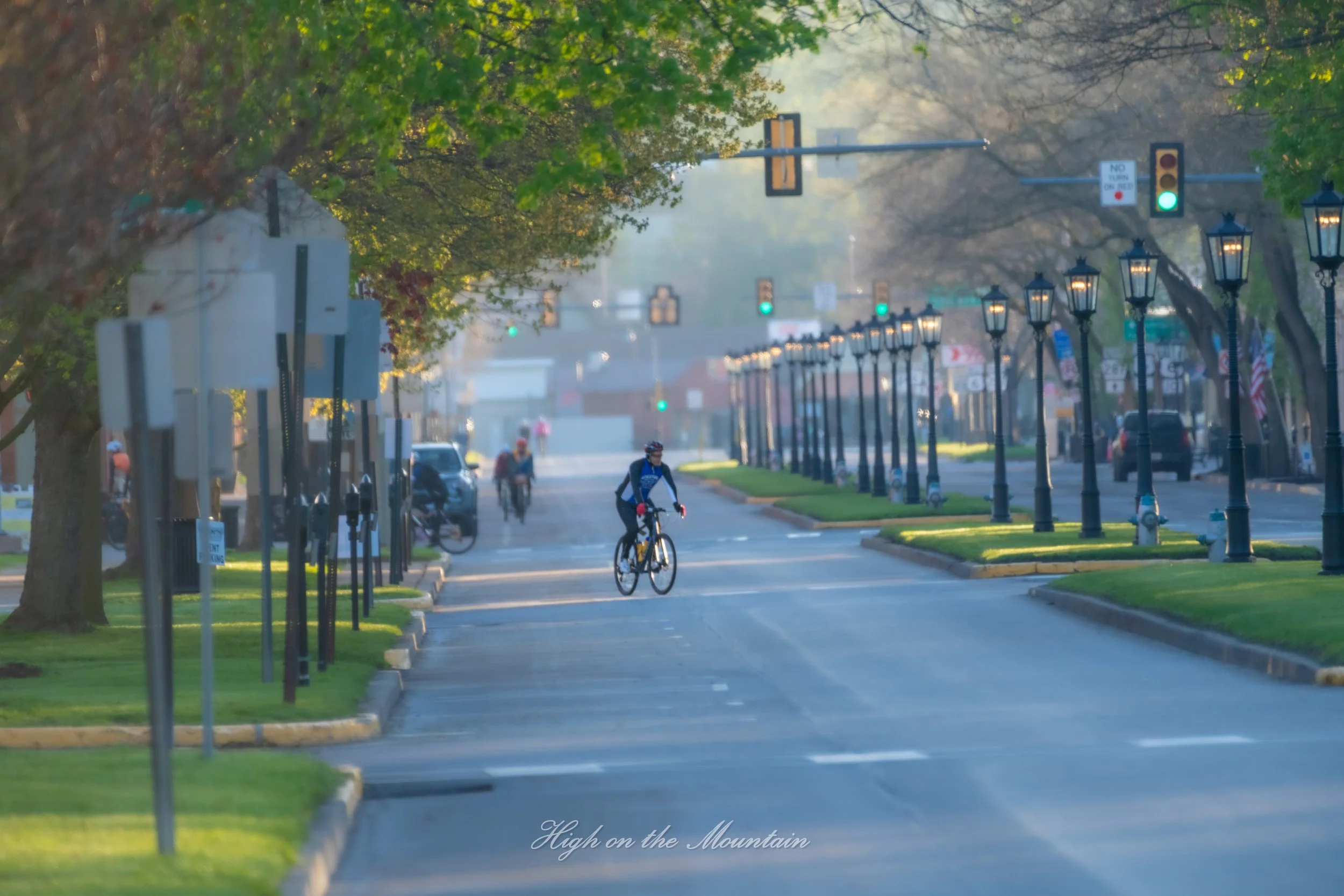 A peaceful street scene with a cyclist riding along a tree-lined road, lantern-style streetlights on the sidewalk, and traffic lights showing green signals in the distance. The street appears to be in a small town or suburban area during early mornin