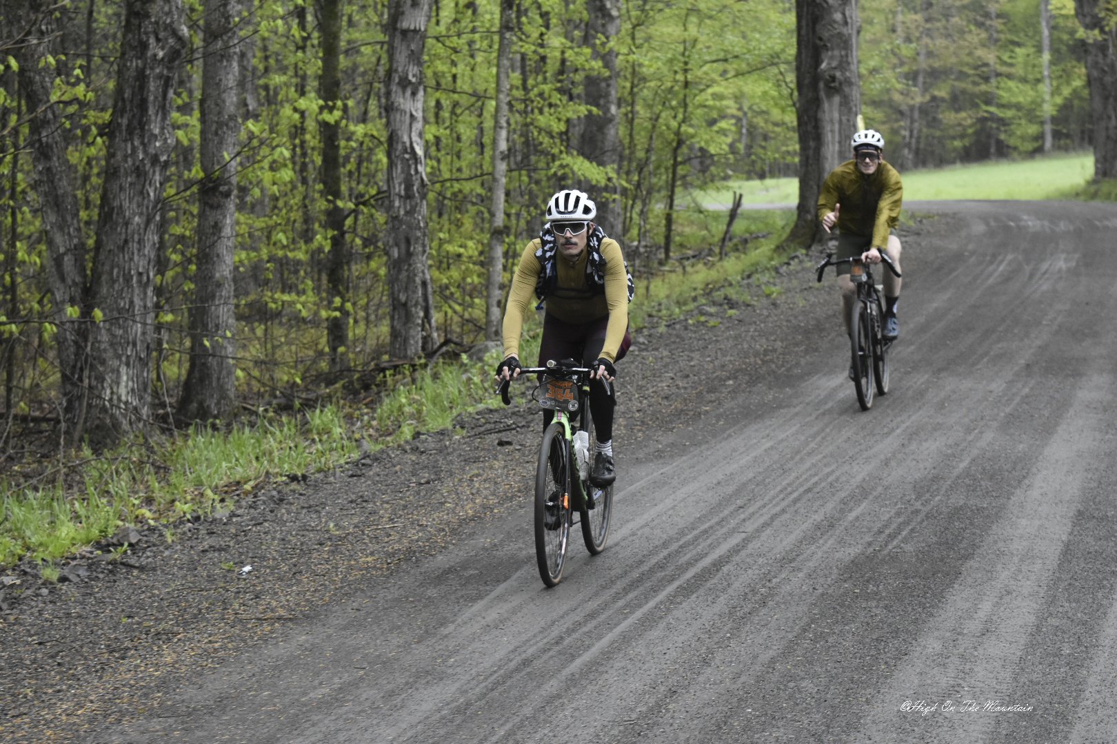 Two men riding mountain bikes on a dirt trail through a green forest. Both wear helmets and sunglasses, with backpacks on their backs.