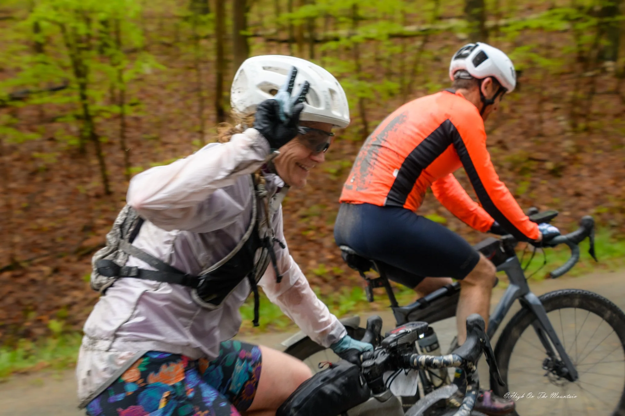 Two people riding mountain bikes on a trail through a wooded area, wearing helmets and outdoor gear