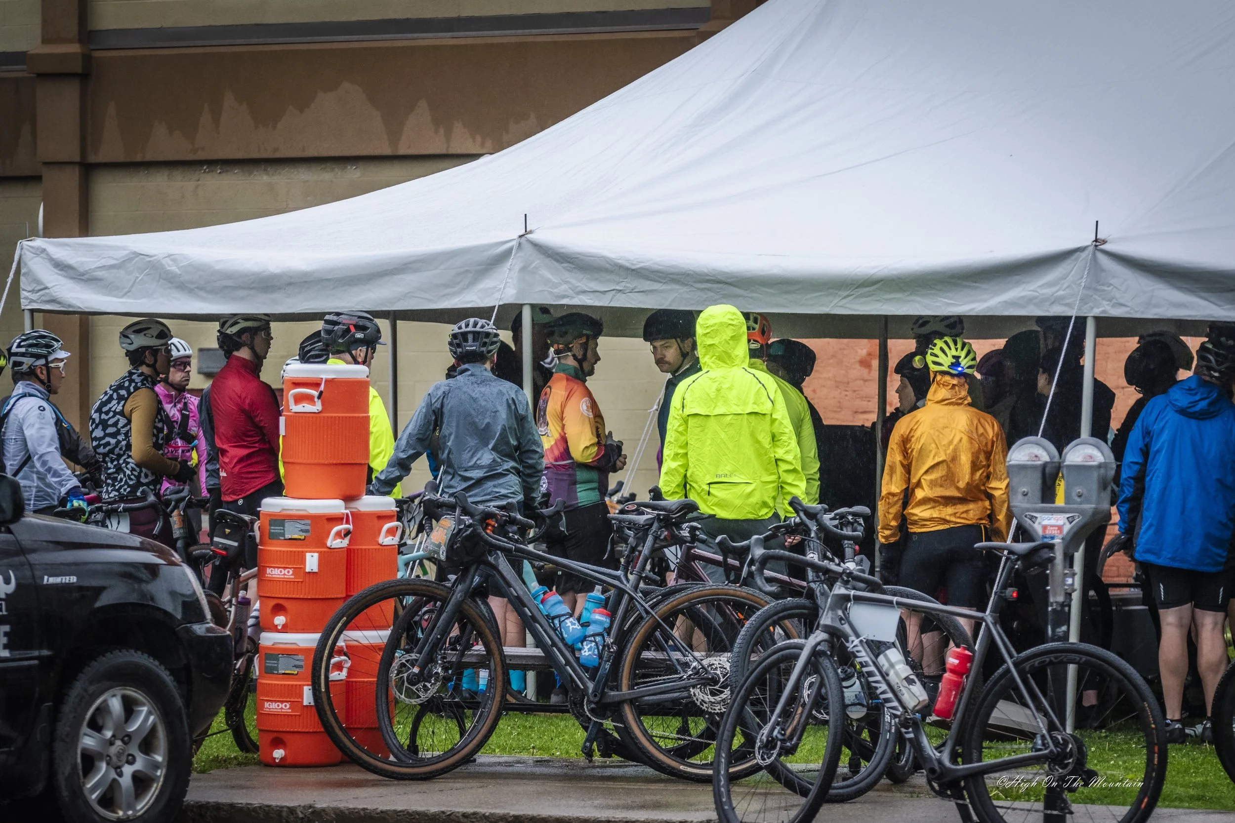 A group of cyclists wearing helmets and rain gear, gathered under a canopy, with some bicycles parked nearby and orange water coolers visible in the foreground.