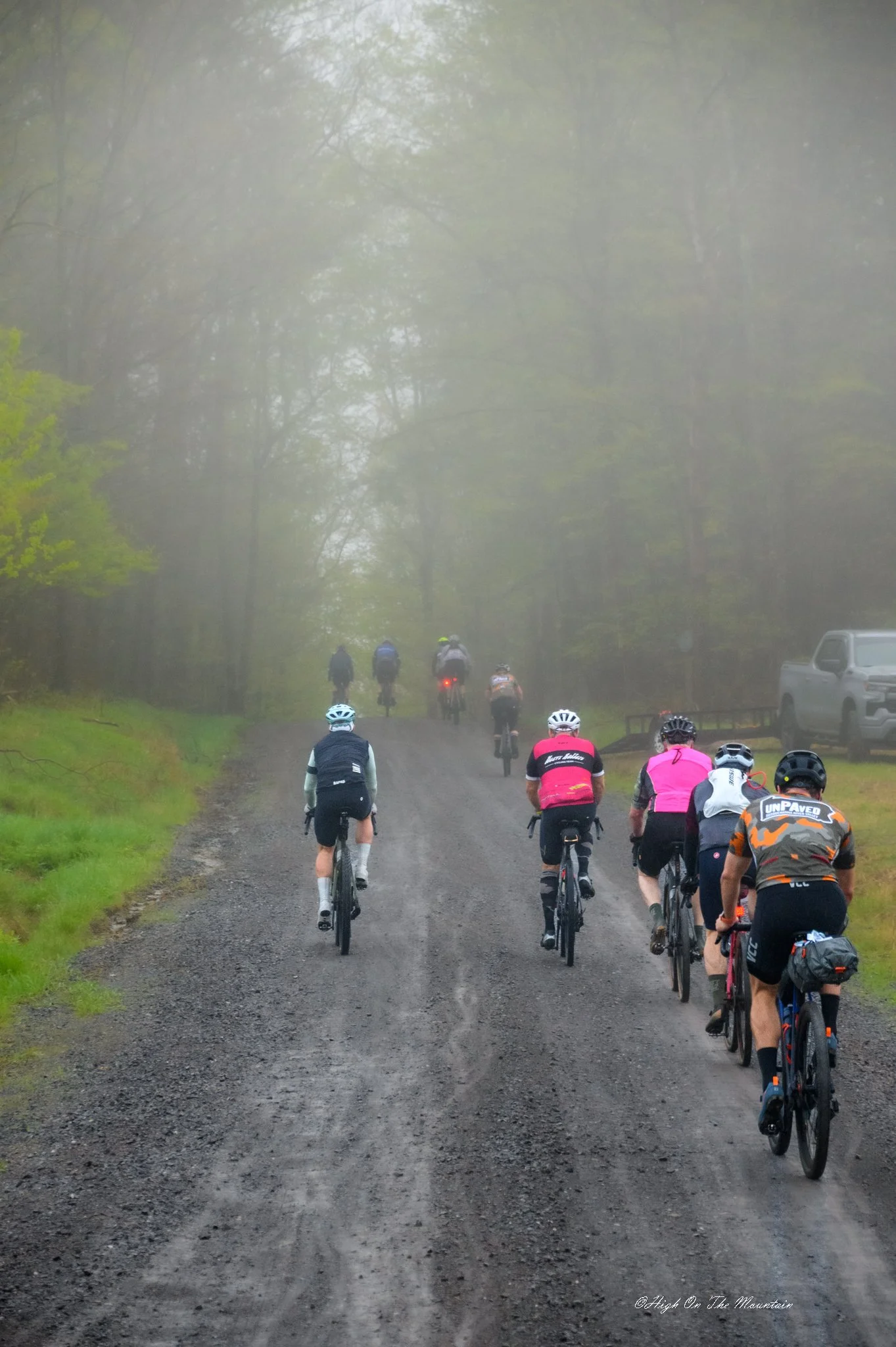A group of cyclists riding on a dirt trail through a foggy forested area, with some in the distance and some closer to the camera. The scene appears to be early morning or late afternoon.