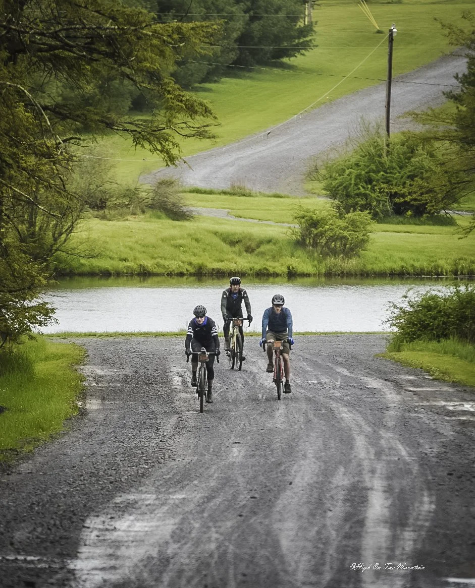 Three bicyclists riding on a gravel path near a lake, surrounded by green trees and grass, with a gravel road and a power pole in the background.