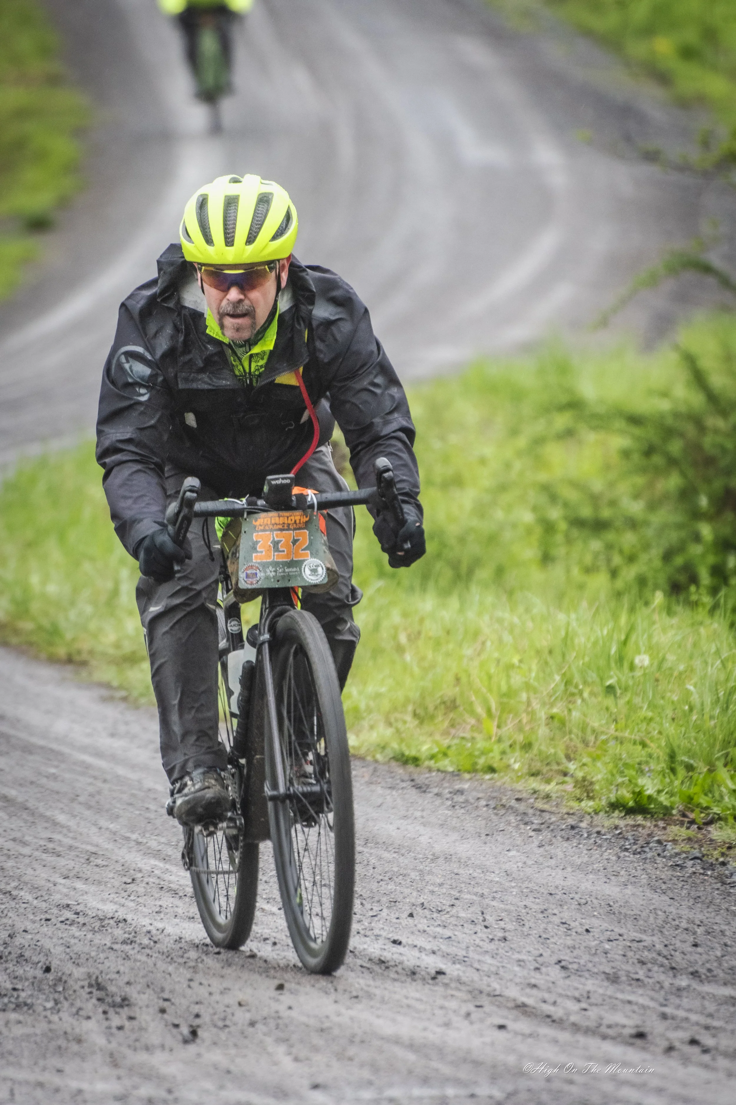 A man in a black jacket and pants riding a mountain bike on a dirt trail in a lush green outdoor setting. He is wearing a yellow helmet, sunglasses, and a race bib number 332, with a determined expression on his face.