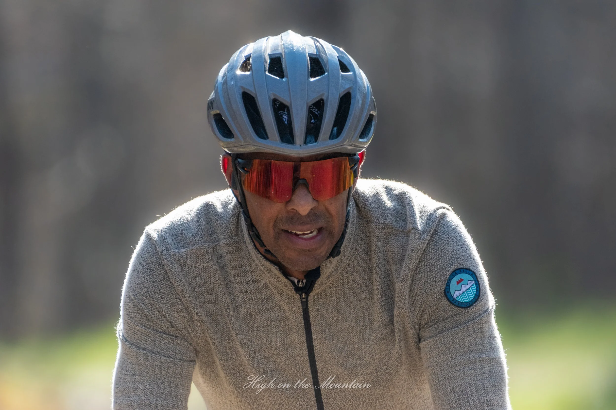A man wearing a gray jacket, red sunglasses, and a white bike helmet with multiple vents, riding outdoors on a sunny day with a blurred, natural background.