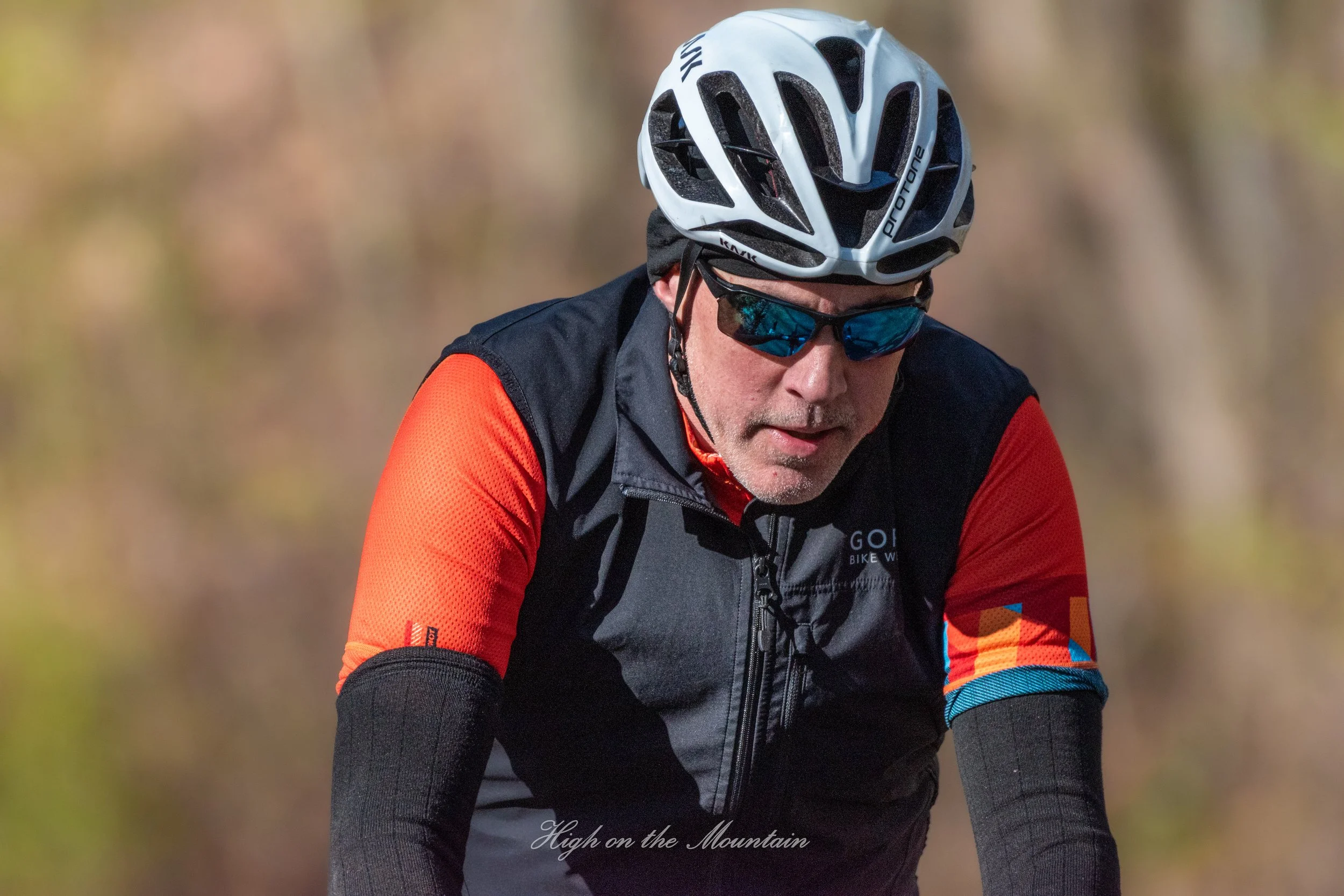 A man wearing a helmet, sunglasses, and a colorful long-sleeve biking jersey riding a bicycle outdoors with a blurred background.