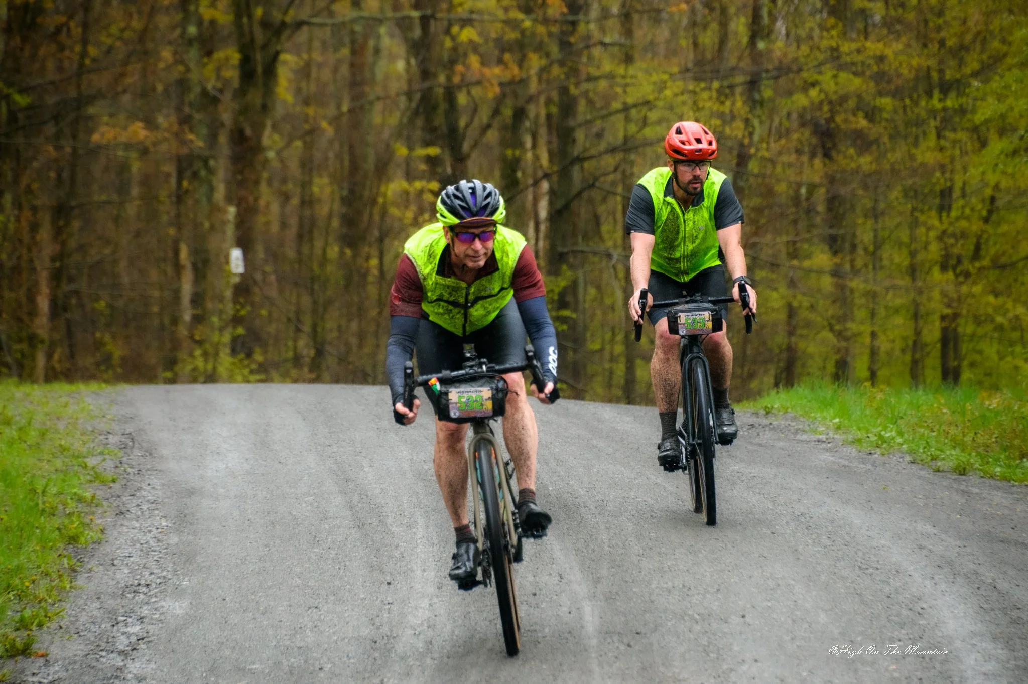 Two mountain bikers riding on a gravel trail through a forest with yellowing leaves, wearing helmets and fluorescent vests.