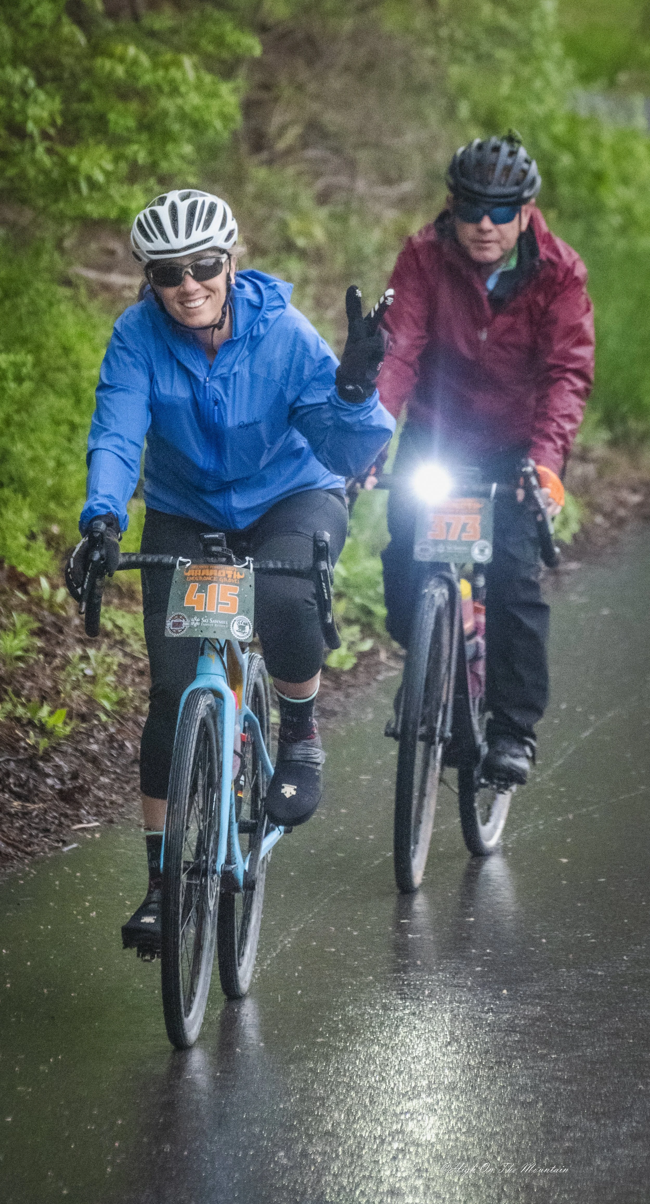 Two cyclists riding on a wet trail in a green, wooded area. One cyclist is smiling and making a peace sign, wearing a blue jacket and white helmet. The other cyclist, wearing a red jacket and black helmet, is also riding alongside.