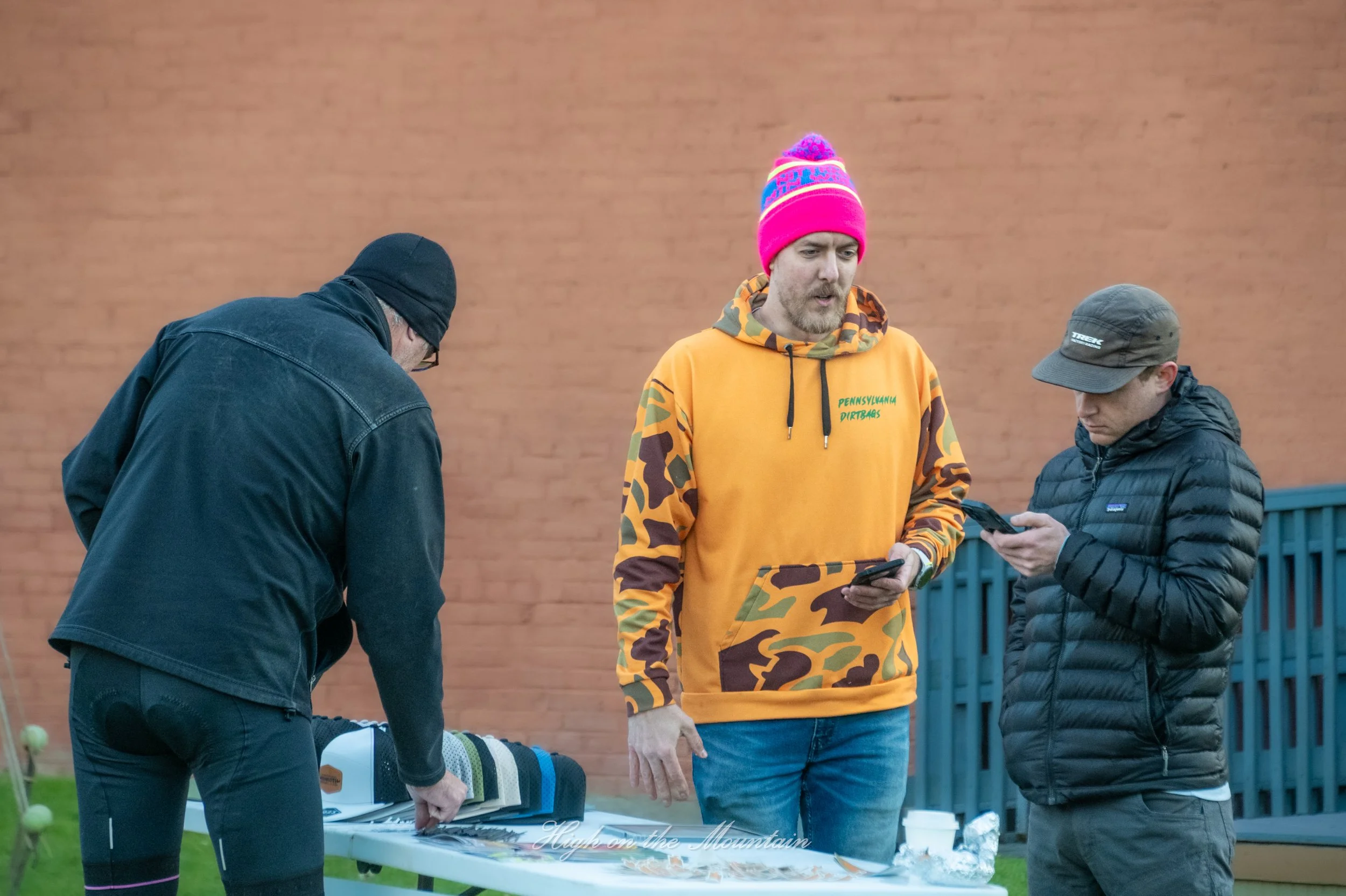 Three men standing around a picnic table outdoors against a brick wall, with one man wearing a bright pink beanie and colorful camouflage hoodie, and the other two men wearing dark clothing and hats, all engaged with their phones or a device on the t