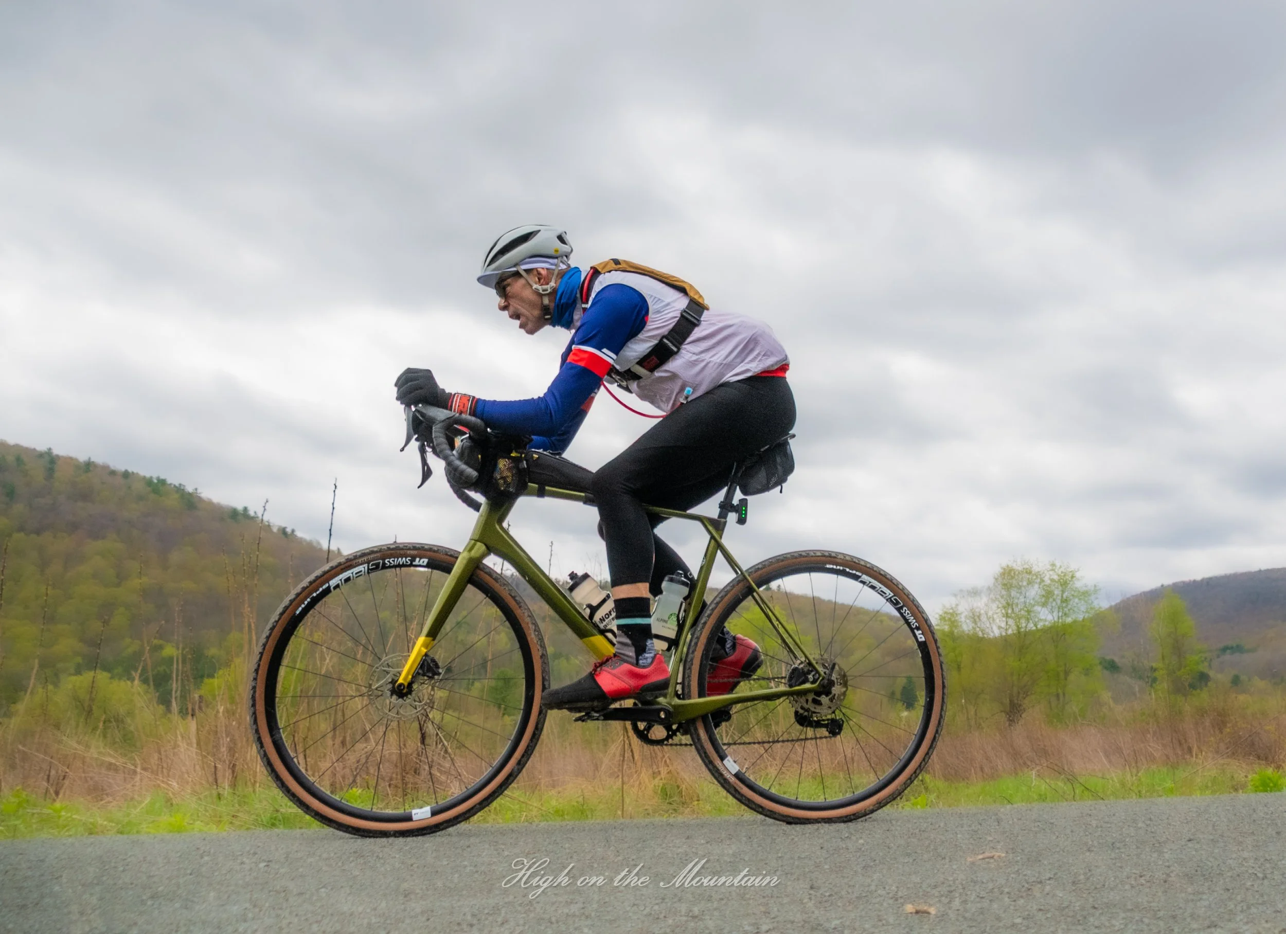Cyclist wearing a white helmet and blue, white, and red cycling outfit riding a mountain bike on a trail with cloudy sky and green hills in the background.