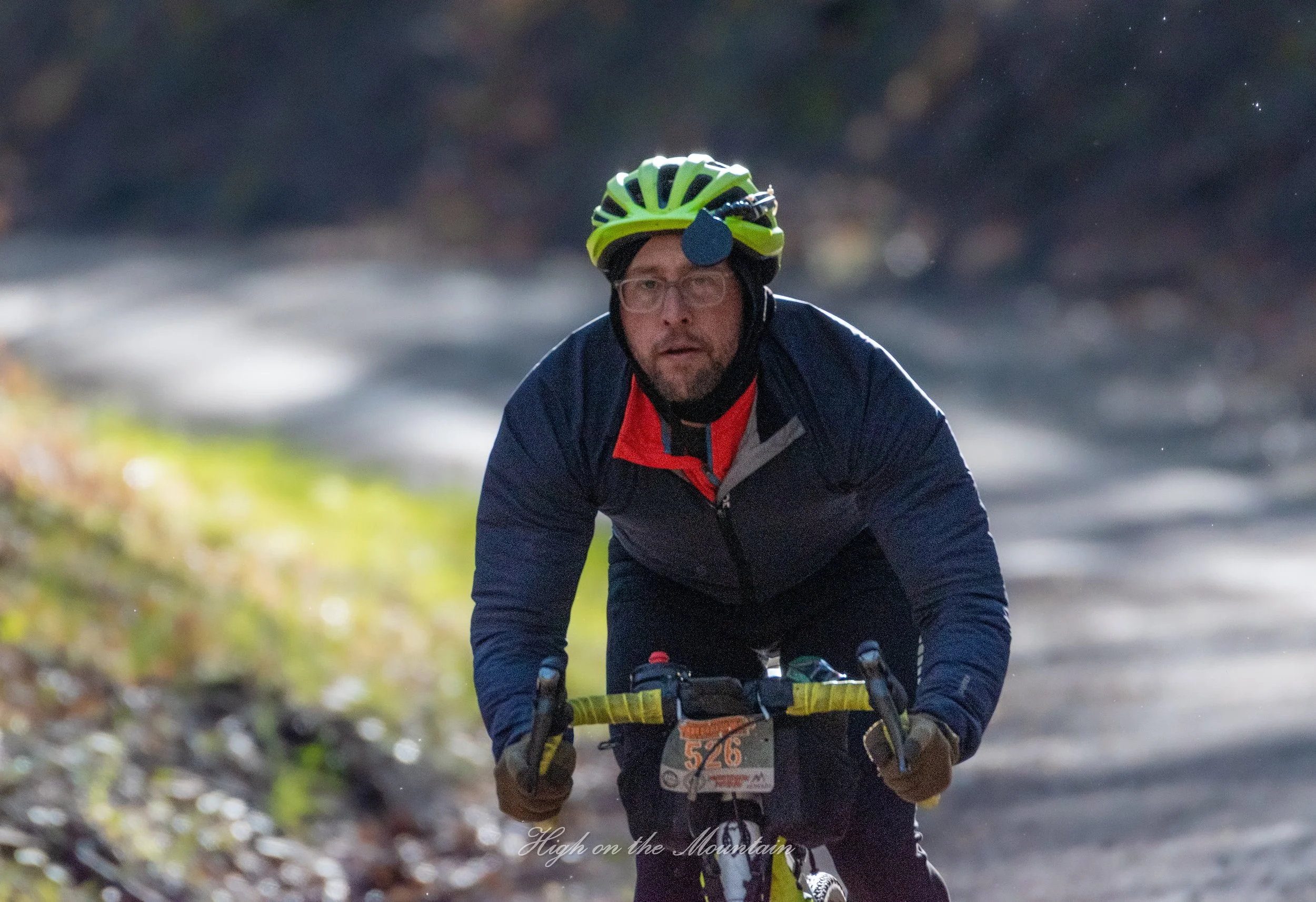 A man riding a mountain bike on a dirt trail in a wooded area. He is wearing a green and black helmet with a headlamp, glasses, a dark jacket, and gloves. He has a focused expression as he navigates the trail.