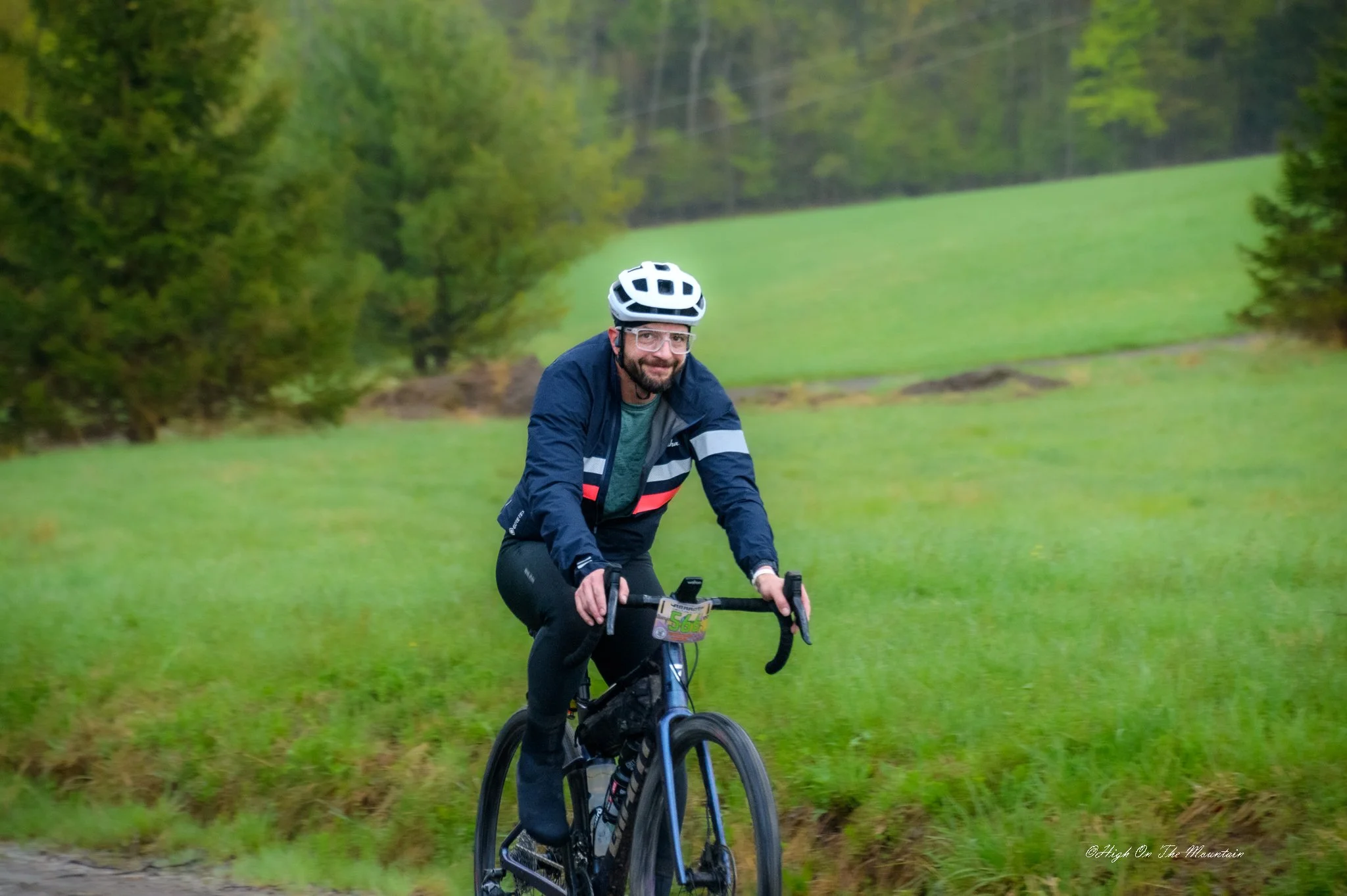 A man with a beard and glasses riding a mountain bike through a green park, wearing a white helmet and a navy and gray jacket.