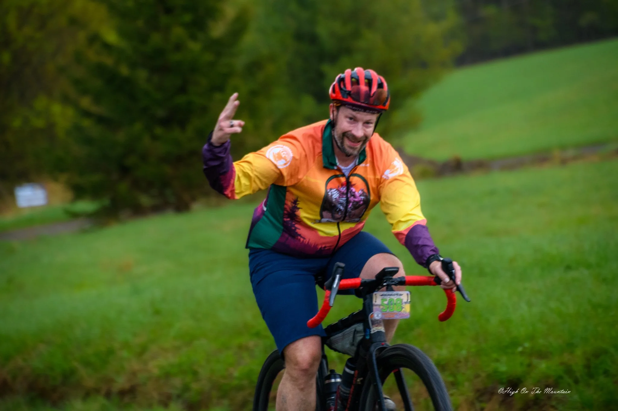 A man riding a mountain bike on a grassy trail, wearing a colorful jacket, helmet, and smiling while pointing to the side.