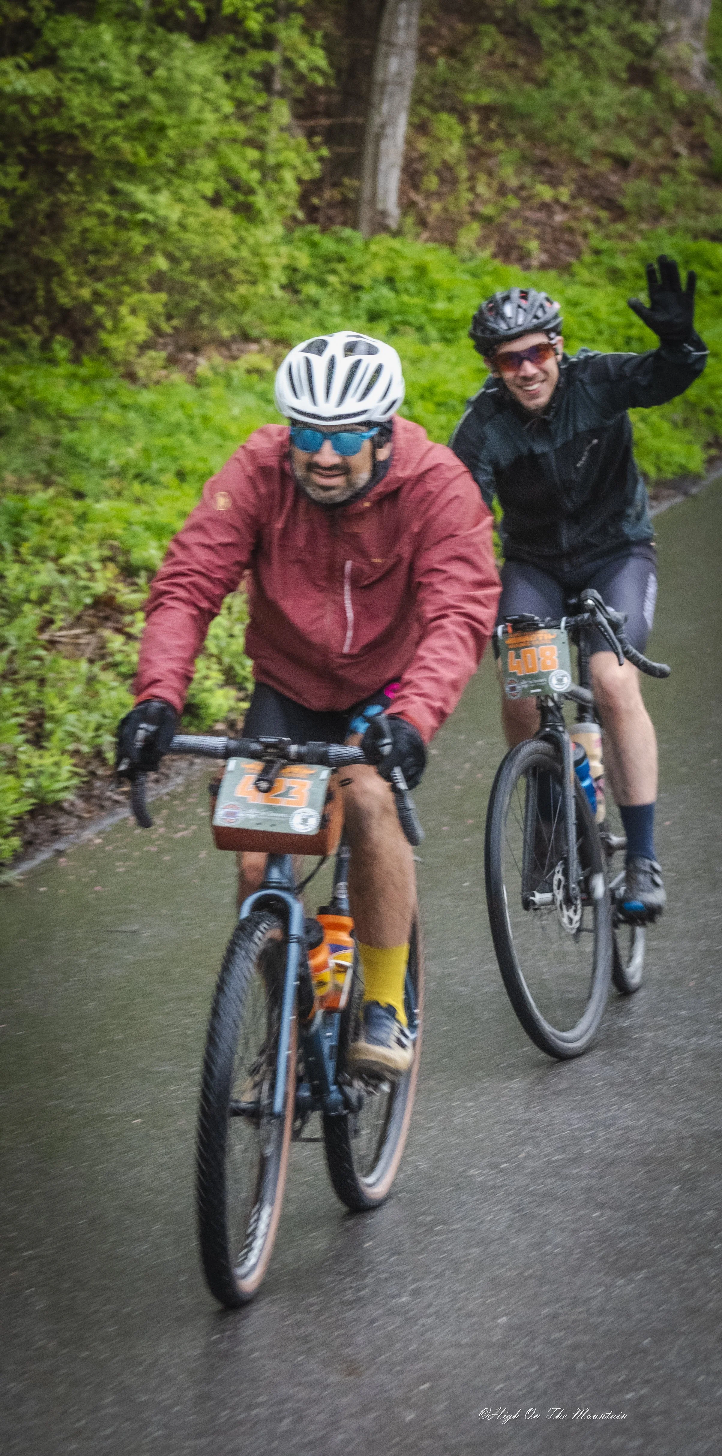 Two men riding bikes on a wet trail surrounded by green trees, wearing helmets and cycling gear, one smiling and waving.
