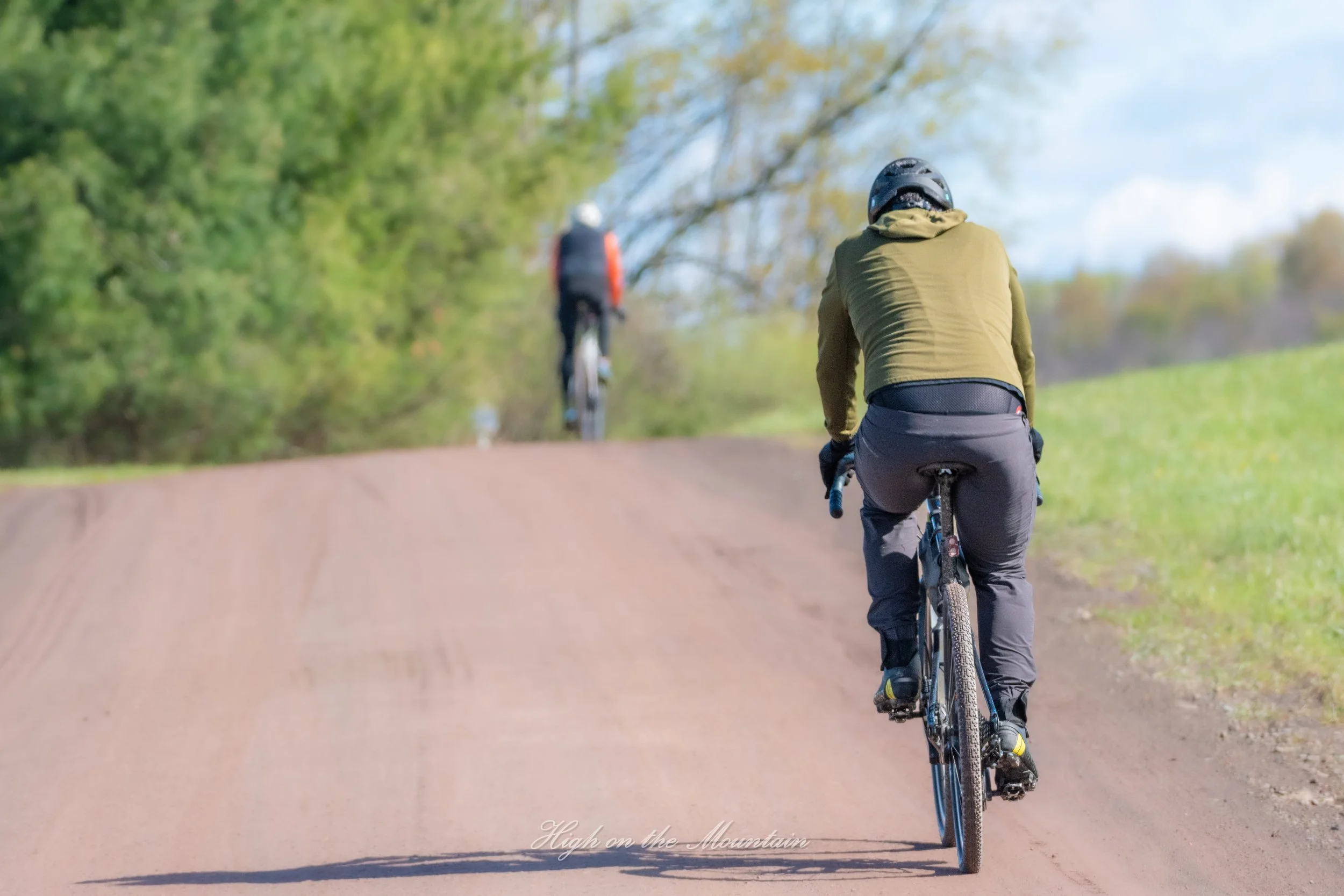 Two cyclists riding on a dirt path surrounded by green trees under a blue sky.