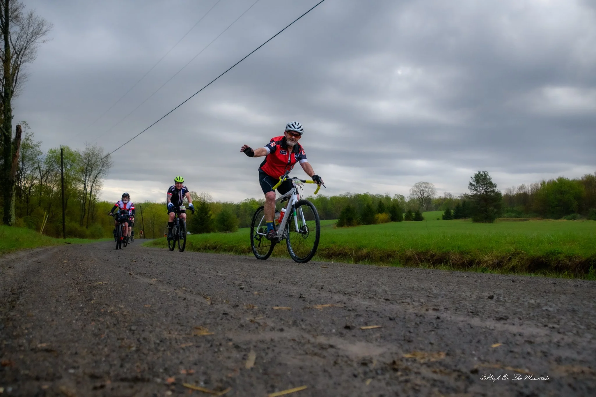 Three cyclists riding on a gravel path in a rural area with green fields and trees under a cloudy sky.